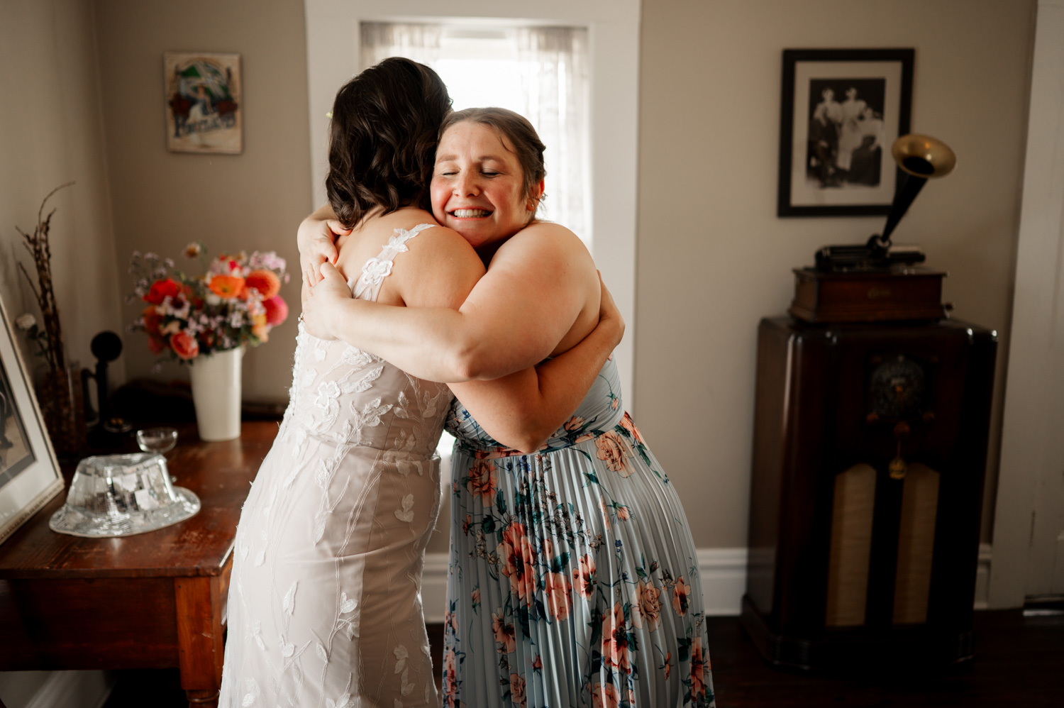 Two women embracing happily in a room with vintage decor and floral accents.