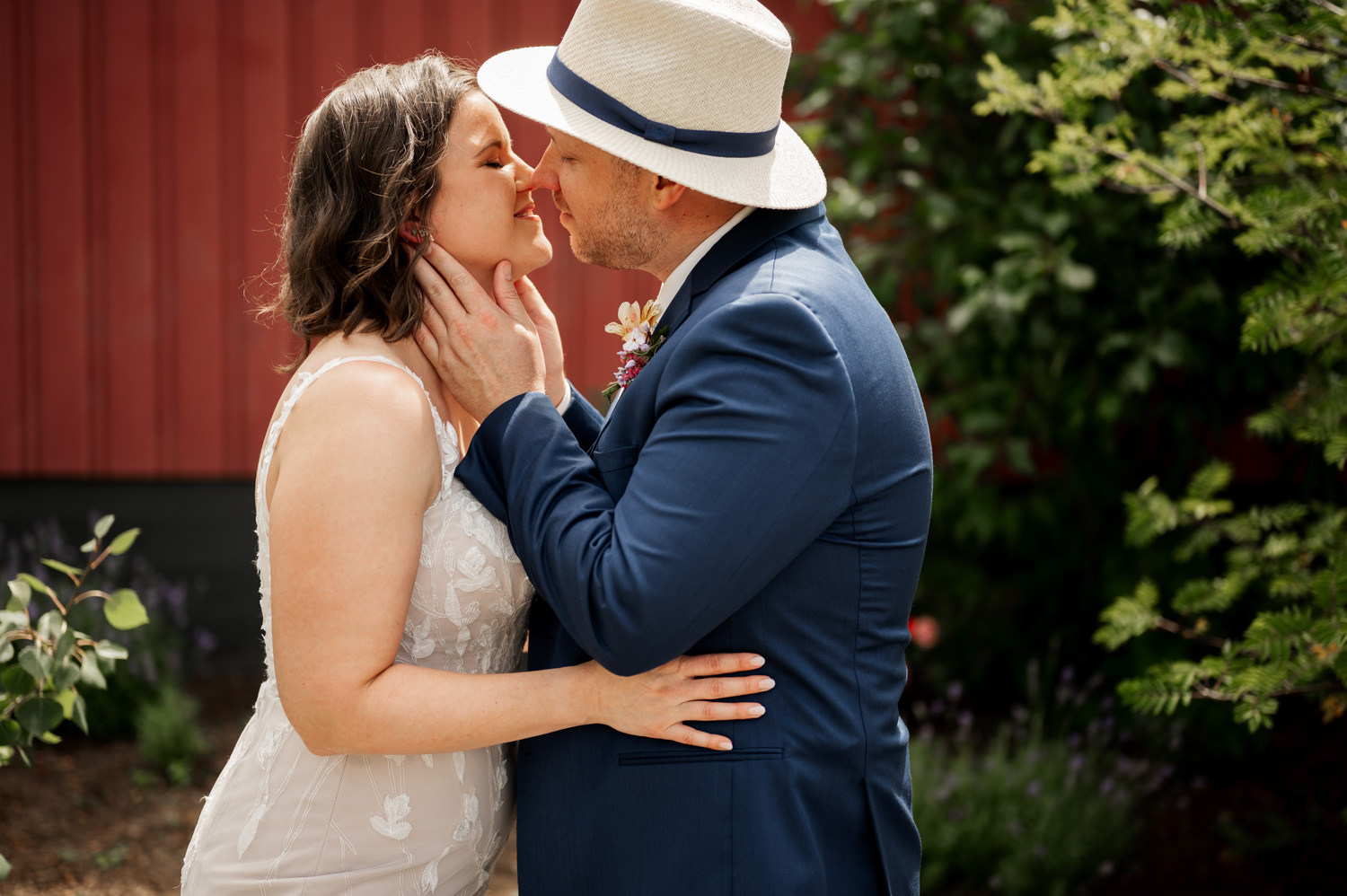 A couple in formal attire shares a tender kiss outdoors at Rossi Farms, with lush greenery and a charming red building visible in the background.