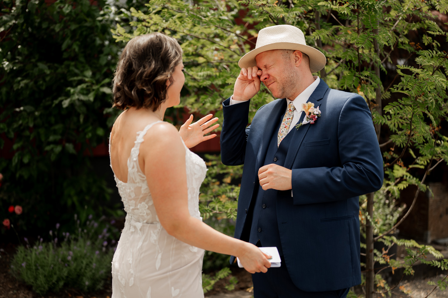 A bride in a white dress gently wipes away her grooms tears as he stands in a blue suit, surrounded by lush greenery at Rossi Farms.