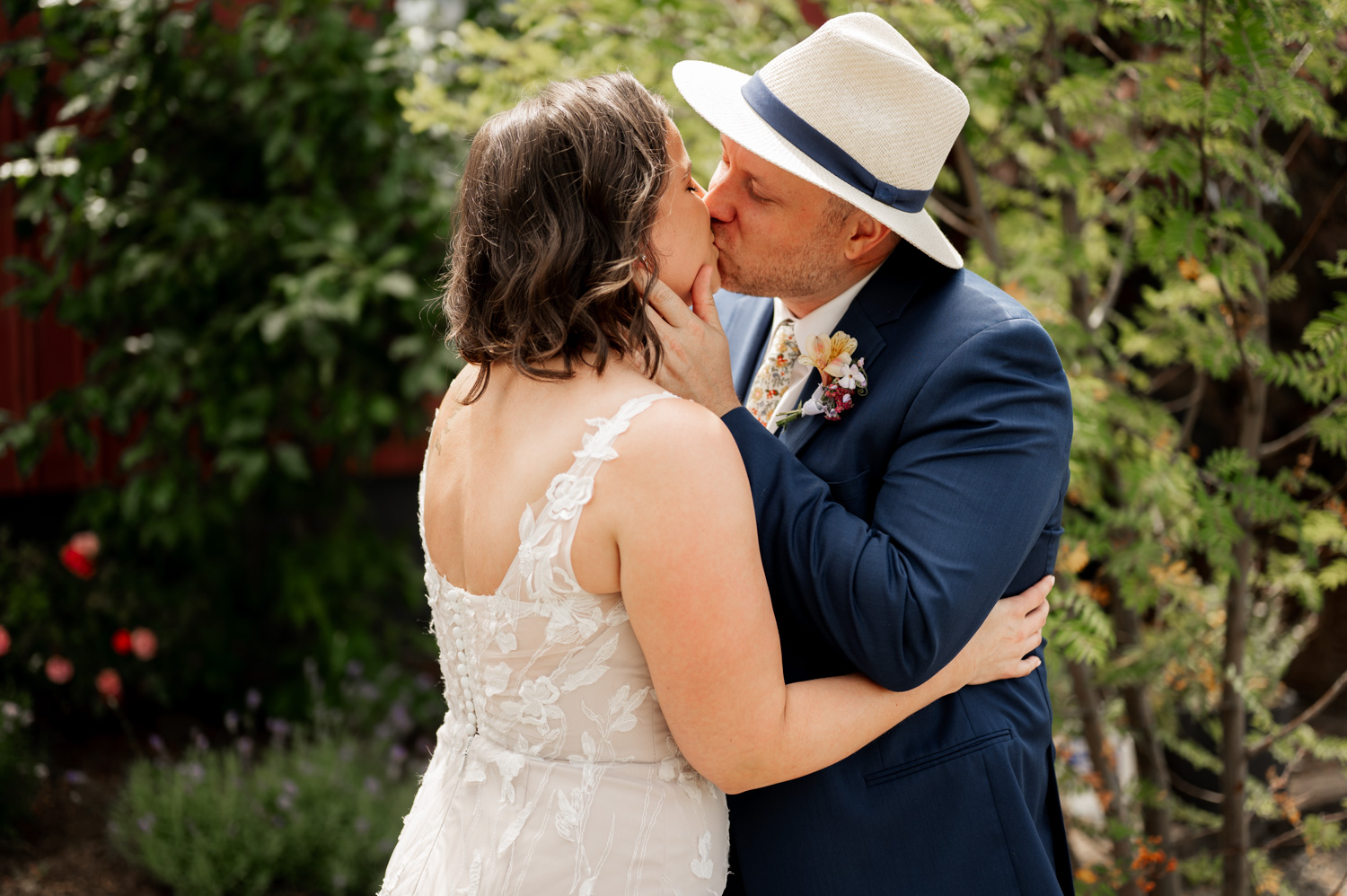 A couple kisses outdoors at Rossi Farms; the woman wears a lace dress, and the man is in a blue suit with a straw hat.