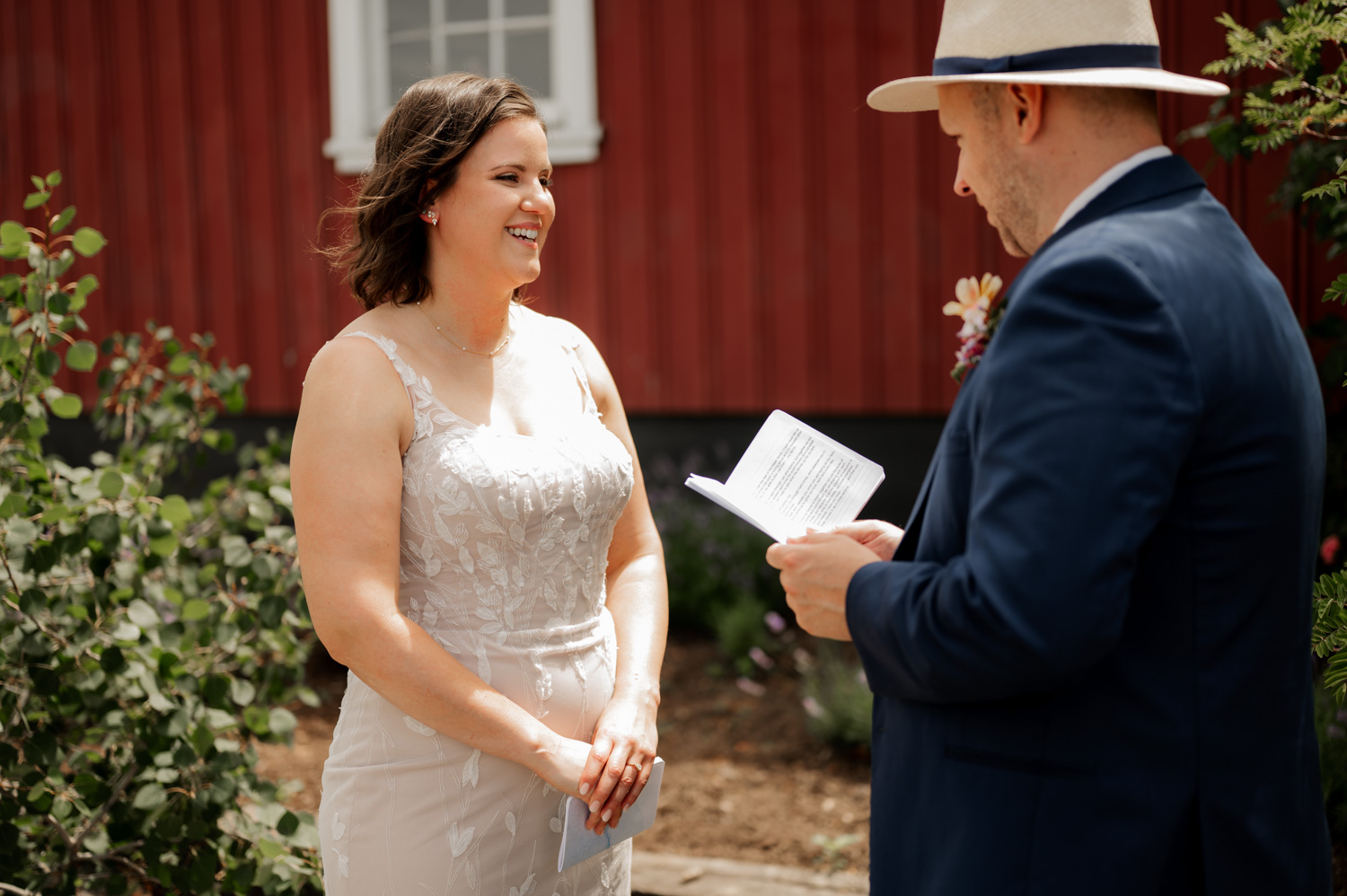 A couple stands outside at Rossi Farms exchanging vows; the woman smiles in a white dress, and the man reads from a book.