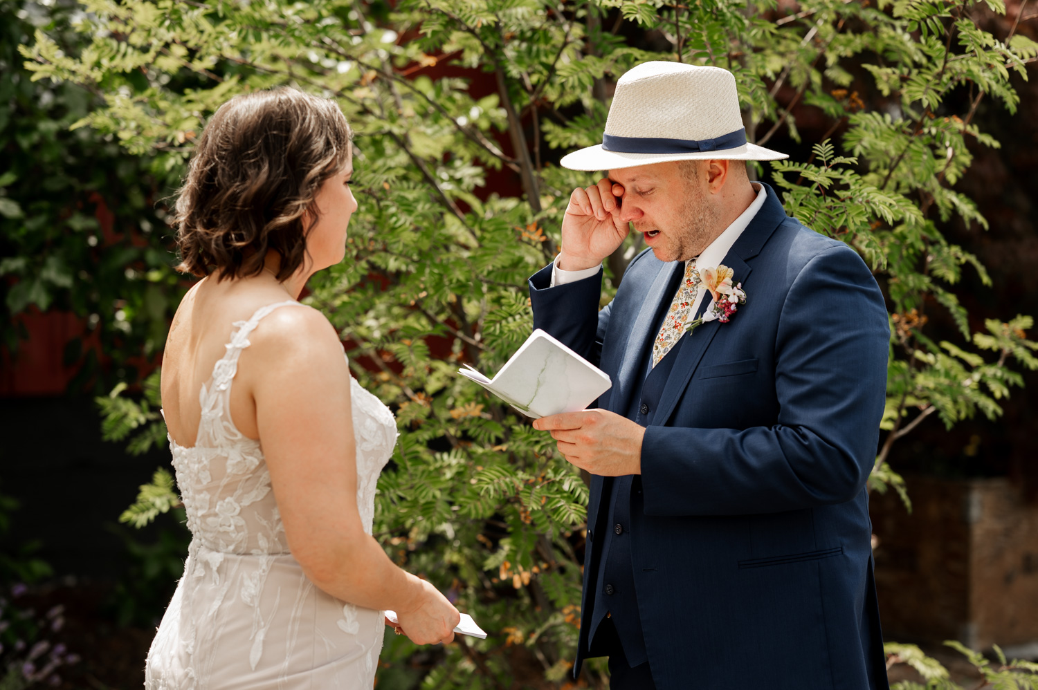 A groom, dressed in a suit, wipes tears from his eyes as he reads vows to his bride. The bride stands opposite him in a beautiful wedding gown. They are surrounded by vibrant green plants and trees at Rossi Farms, creating a picturesque outdoor setting for the ceremony.
