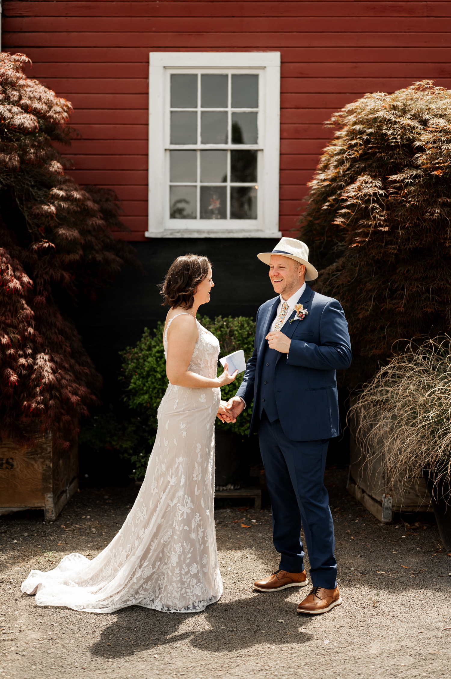 A bride and groom smile at each other, standing in front of a red building with lush plants on either side.