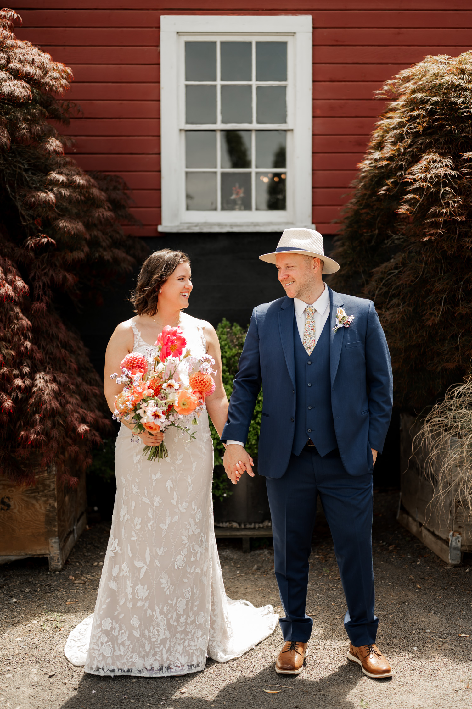 At Rossi Farms, a bride in a white gown holds a bouquet standing beside her groom in a blue suit and hat. They are framed against the backdrop of a vibrant red building.