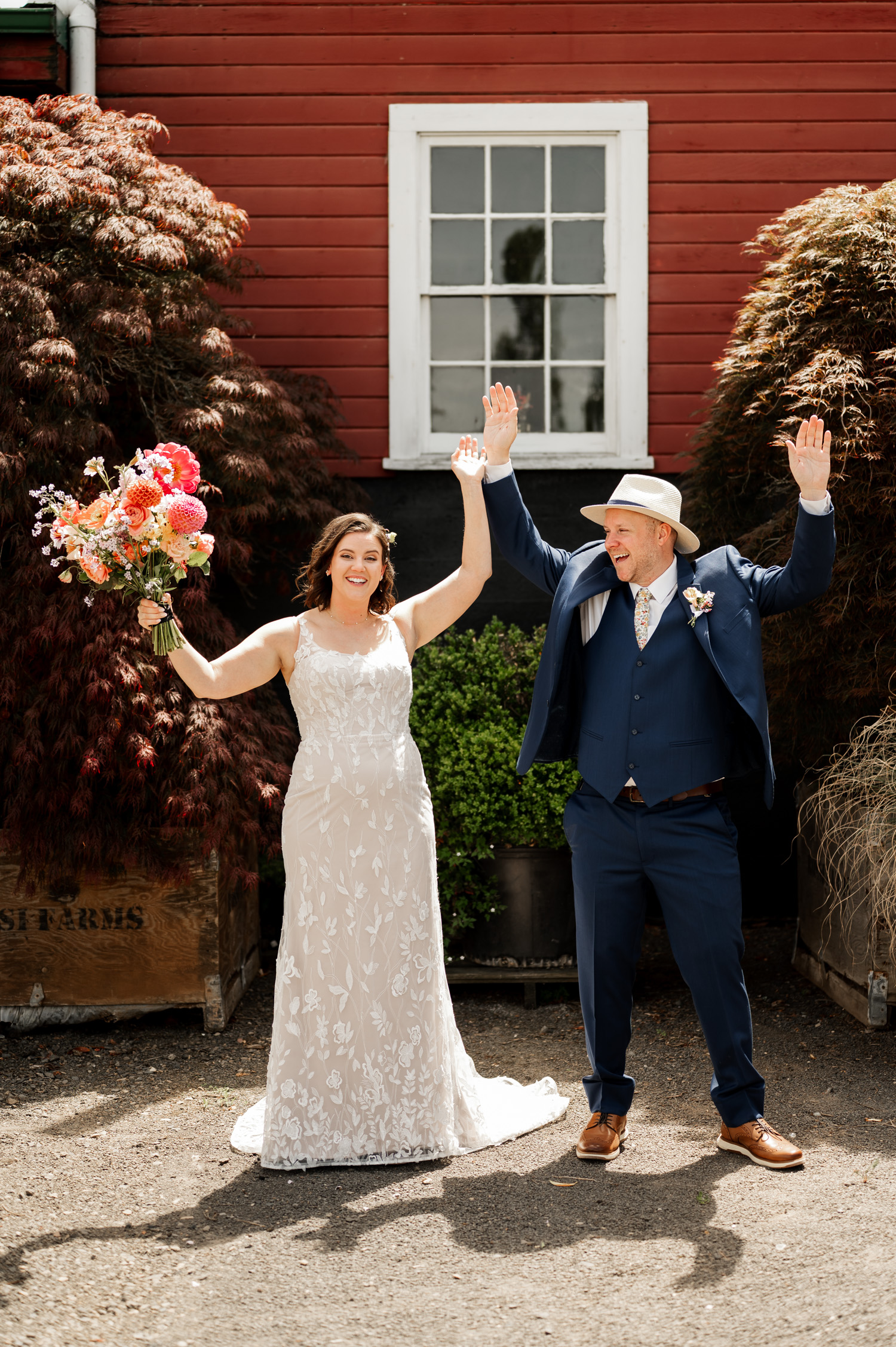 A bride and groom stand happily in front of a vibrant red building at Rossi Farms, with the bride holding a colorful bouquet.