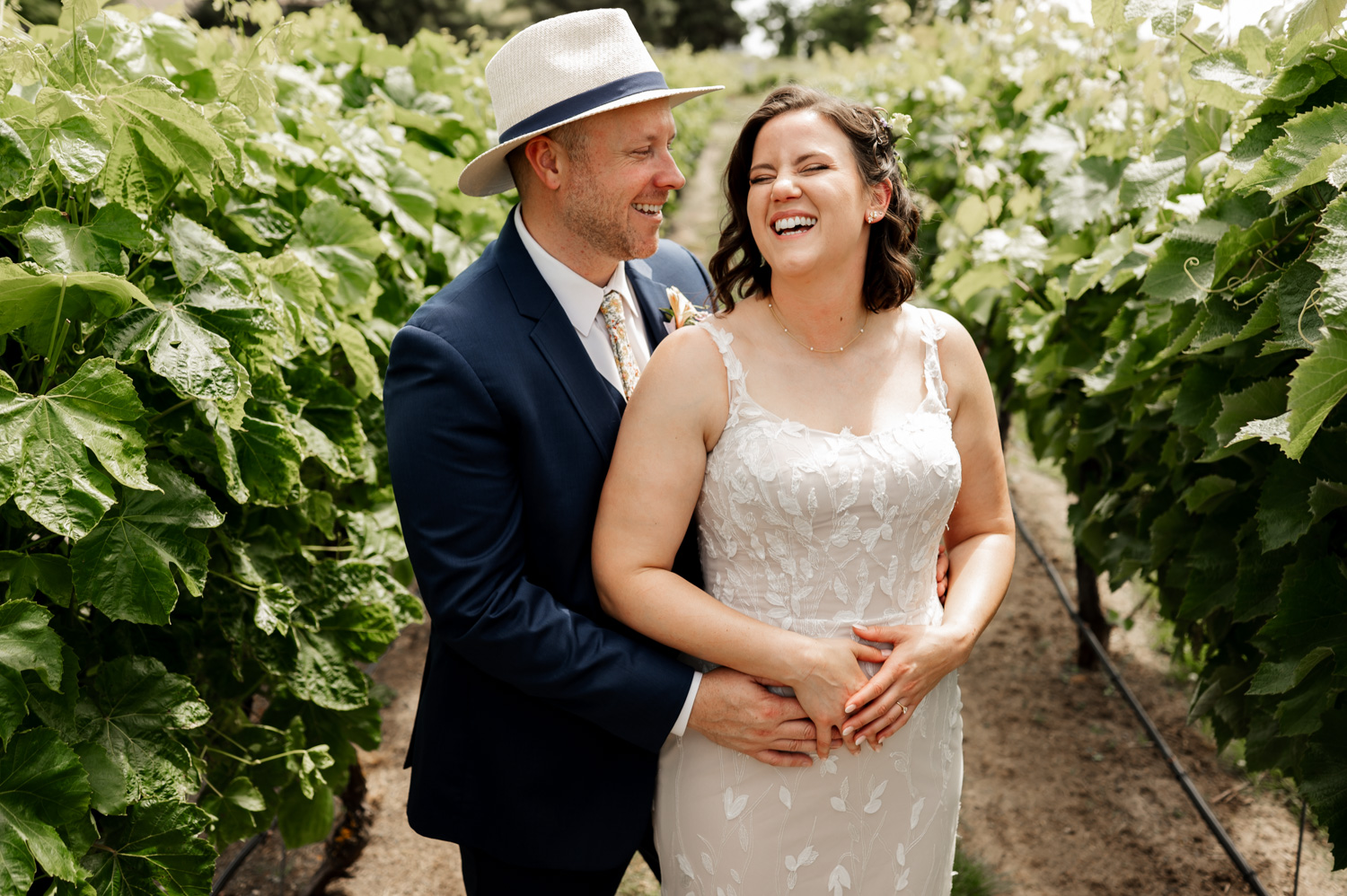 A couple in wedding attire laughing together while standing in a vineyard at Rossi Farms.