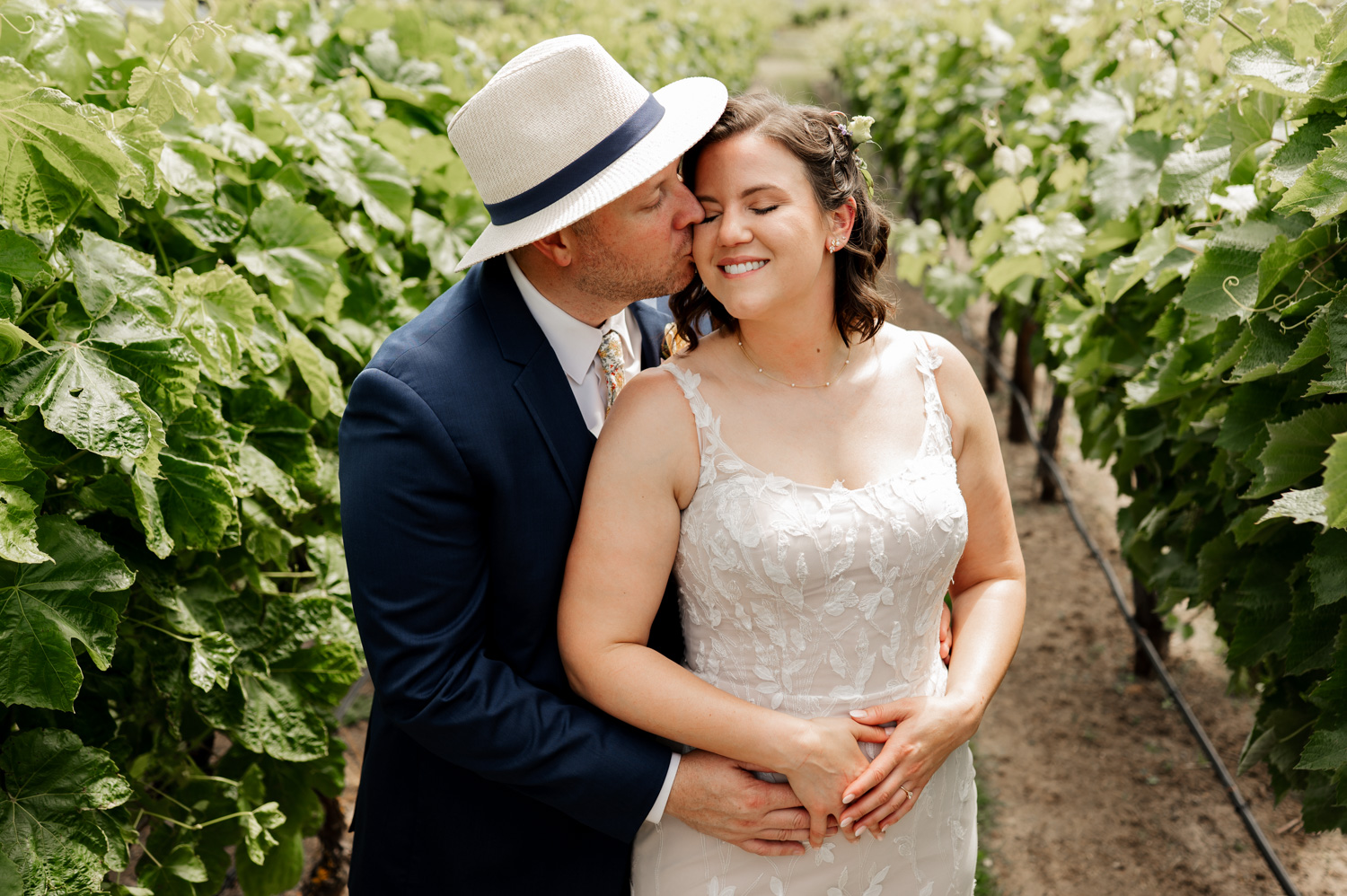 A groom in a suit gently kisses his smiling bride in a white dress amidst rows of lush vineyards at Rossi Farms.