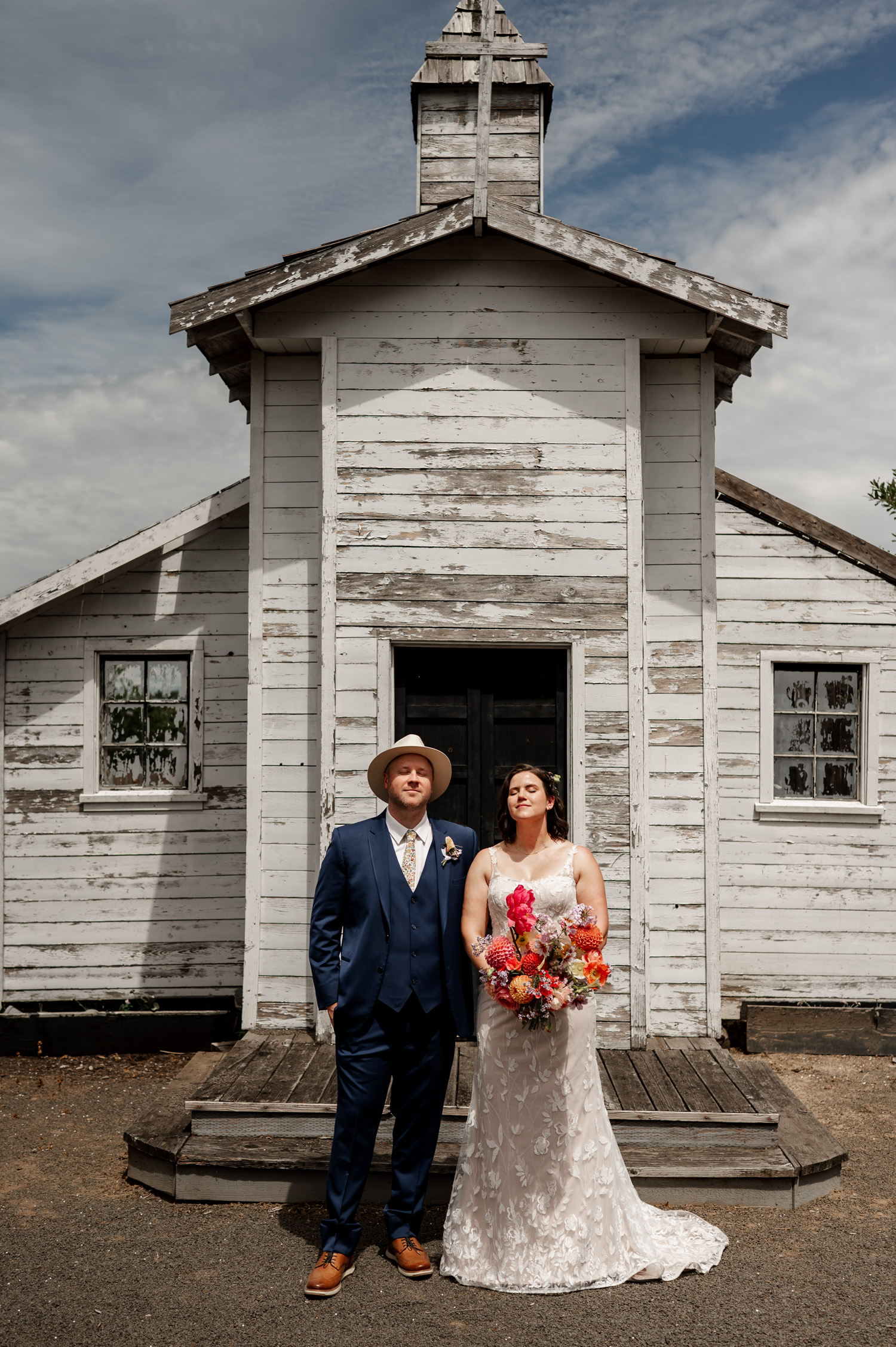 A bride and groom pose with colorful flowers in front of a small rustic church, celebrating their wedding at Rossi Farms.