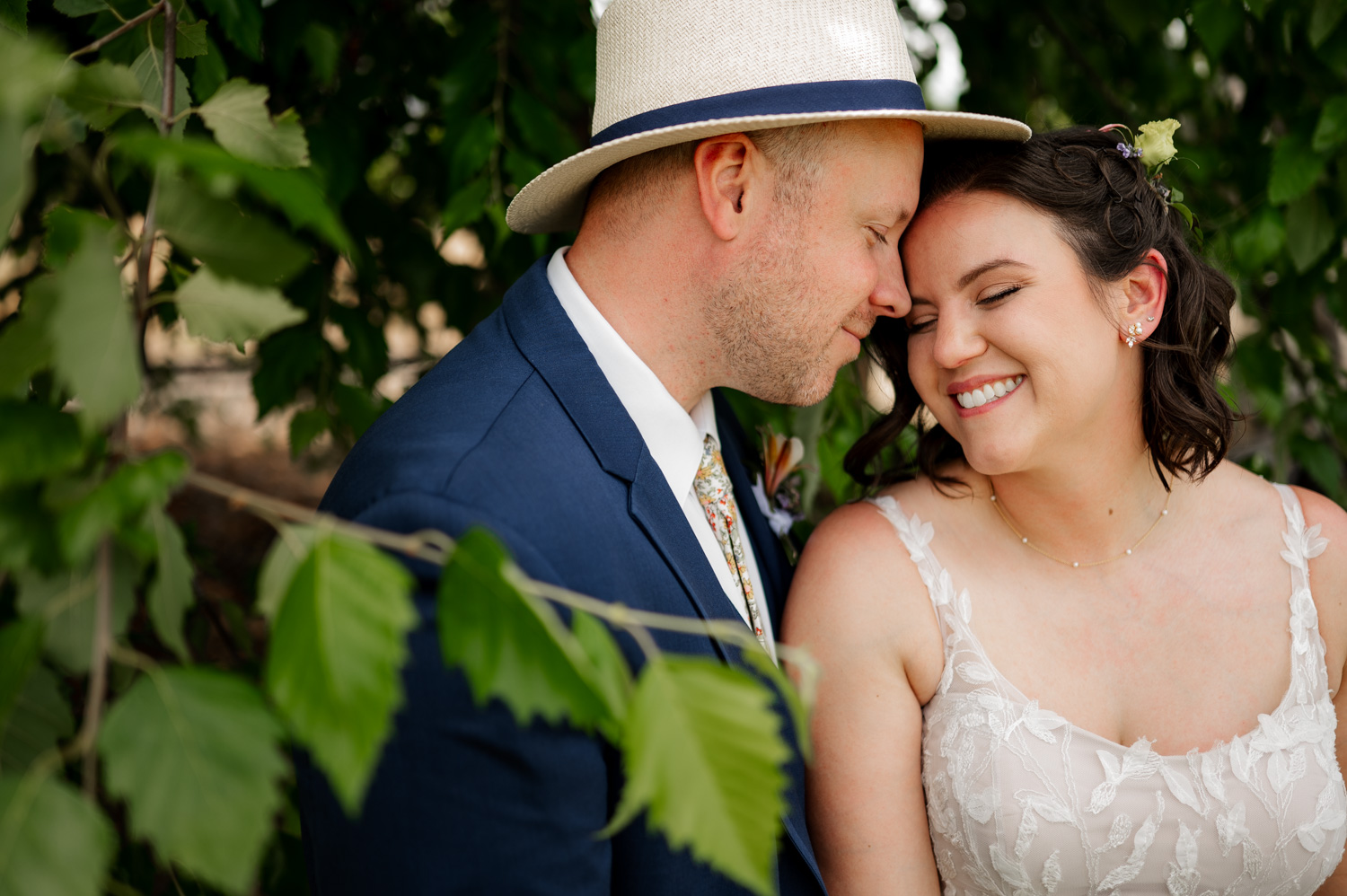 Bride and groom smiling, touching foreheads under leafy branches at Rossi Farms; groom wearing a hat and blue suit.