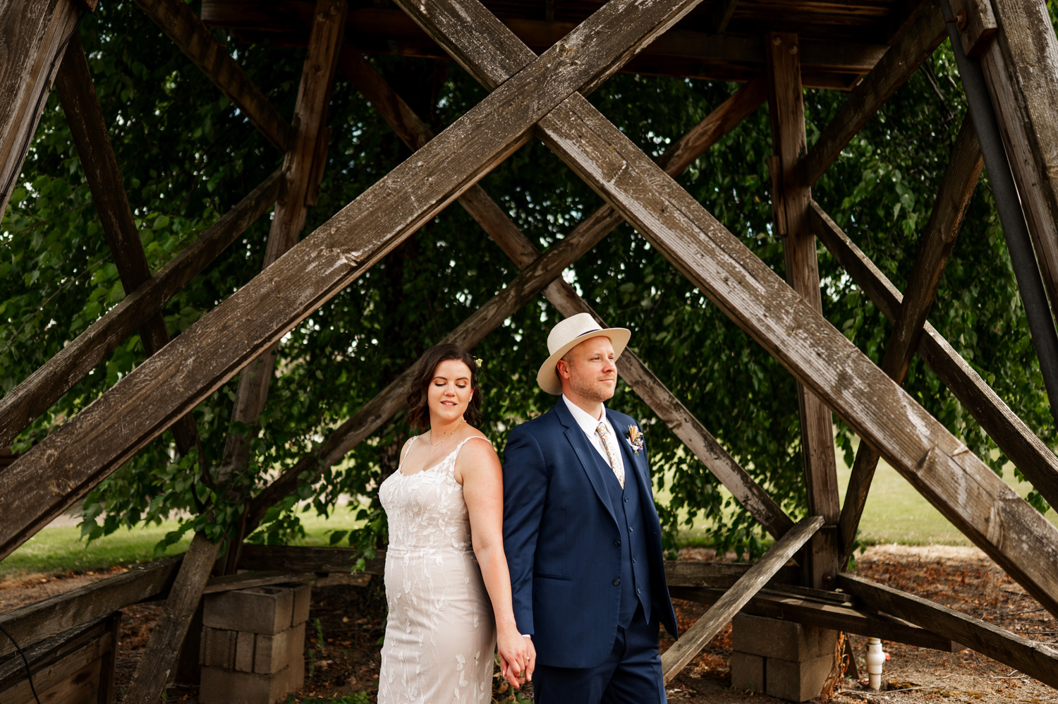 A couple in elegant attire holds hands under a wooden structure, surrounded by greenery at Rossi Farms.