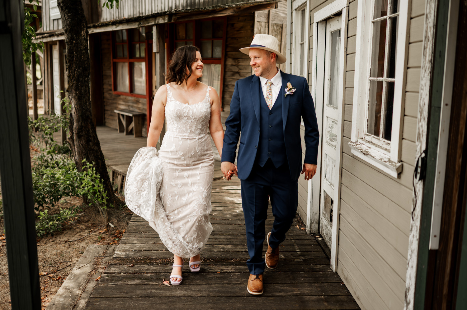 A bride in a lace gown and a groom in a blue suit with a hat hold hands and smile while walking along a wooden path at Rossi Farms.