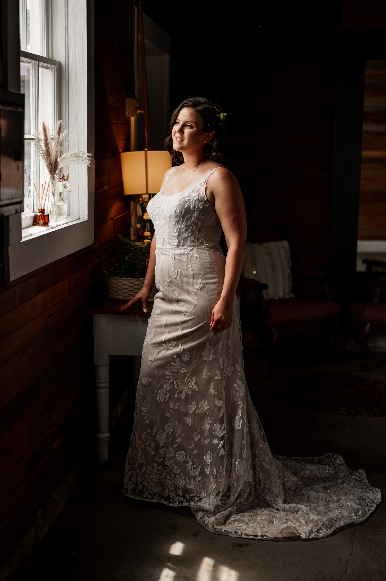 A bride in an embroidered gown stands by a window in a warmly lit room at Rossi Farms, looking outside thoughtfully.