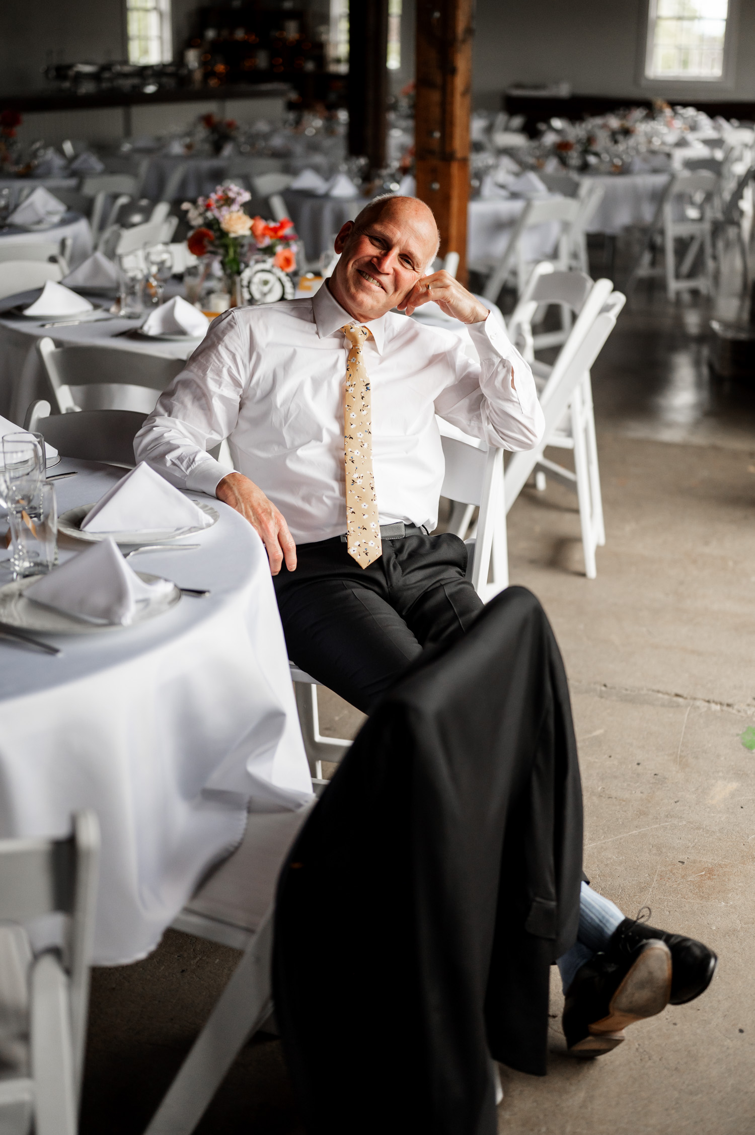 A man in formal attire smiling while seated at a banquet table with white tablecloths and floral centerpieces; his jacket draped over the chair.