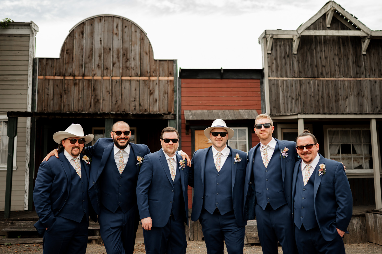 Six groomsmen in blue suits and sunglasses laugh together at Rossi Farms, with rustic buildings in the background.
