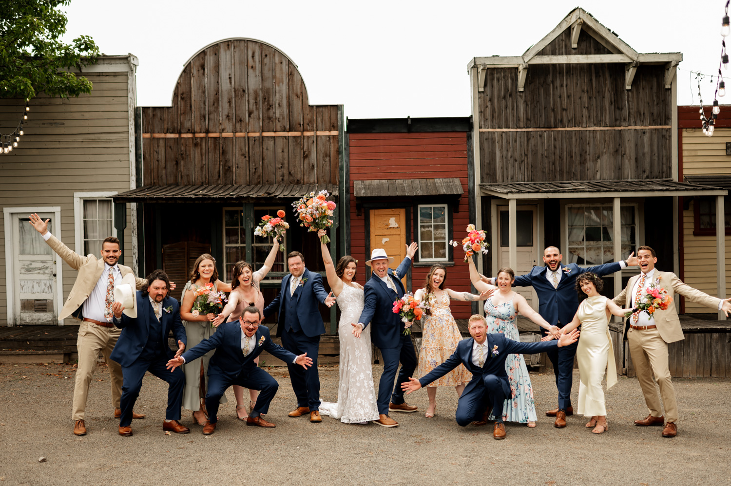The wedding party stands together, smiling in front of rustic buildings at Rossi Farms, dressed in elegant suits and dresses and holding vibrant bouquets.
