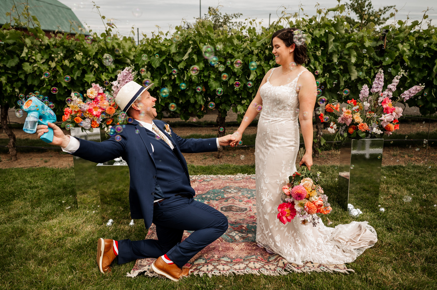 A bride and groom stand at Rossi Farms in a vineyard, surrounded by vibrant flowers and bubbles, both smiling and holding hands.