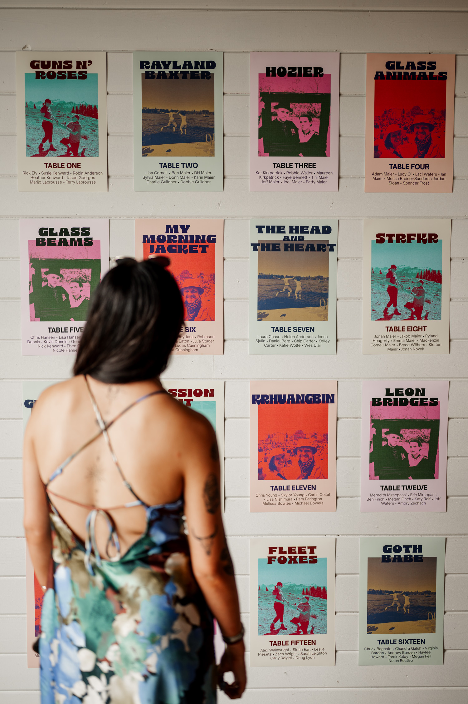 A person in a floral dress stands looking at a wall covered with vibrant posters displaying band names, table numbers, and wedding photos from Rossi Farms.
