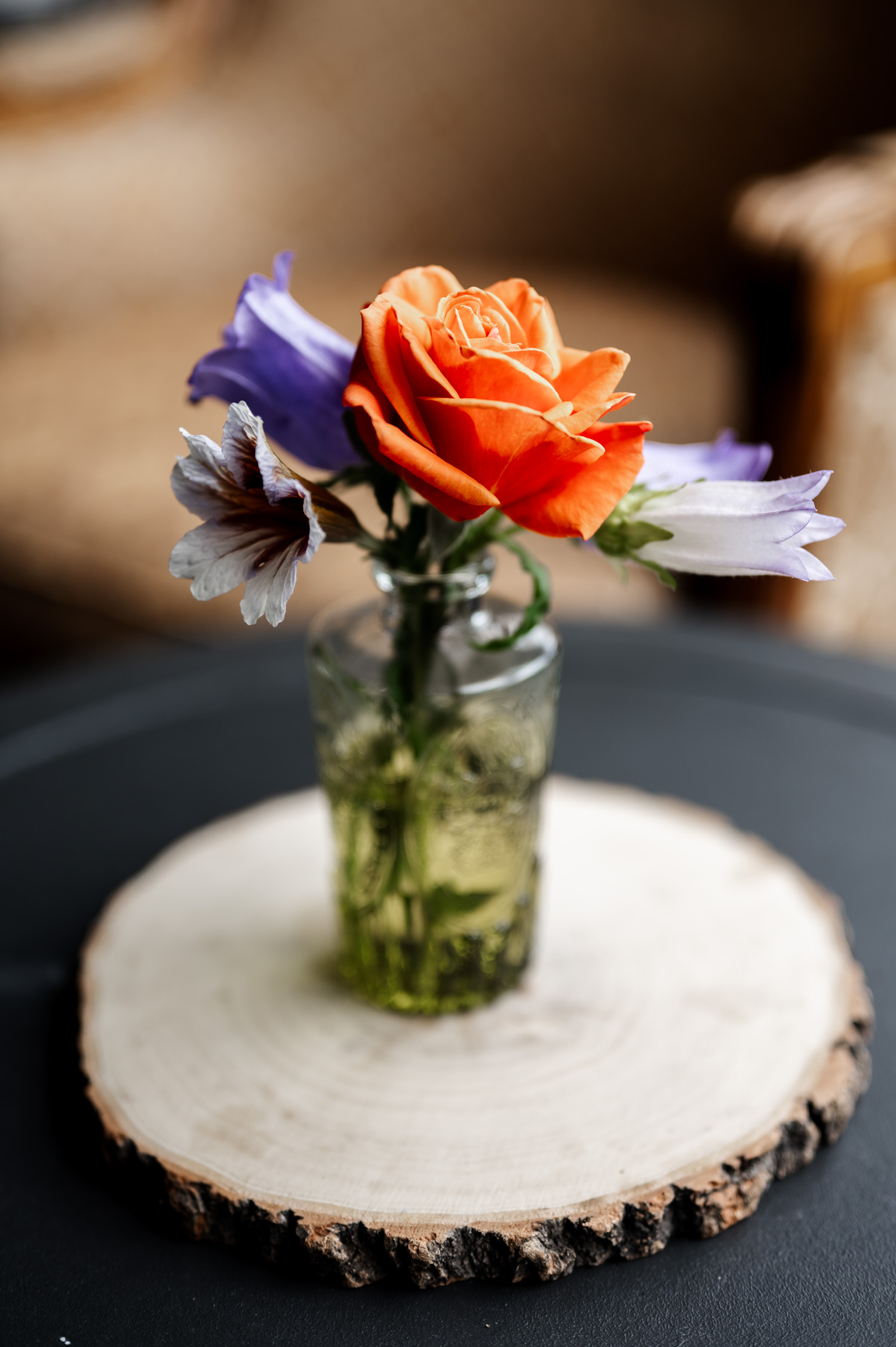 An orange rose and purple flowers are elegantly arranged in a glass vase, placed on a wooden slab centerpiece.