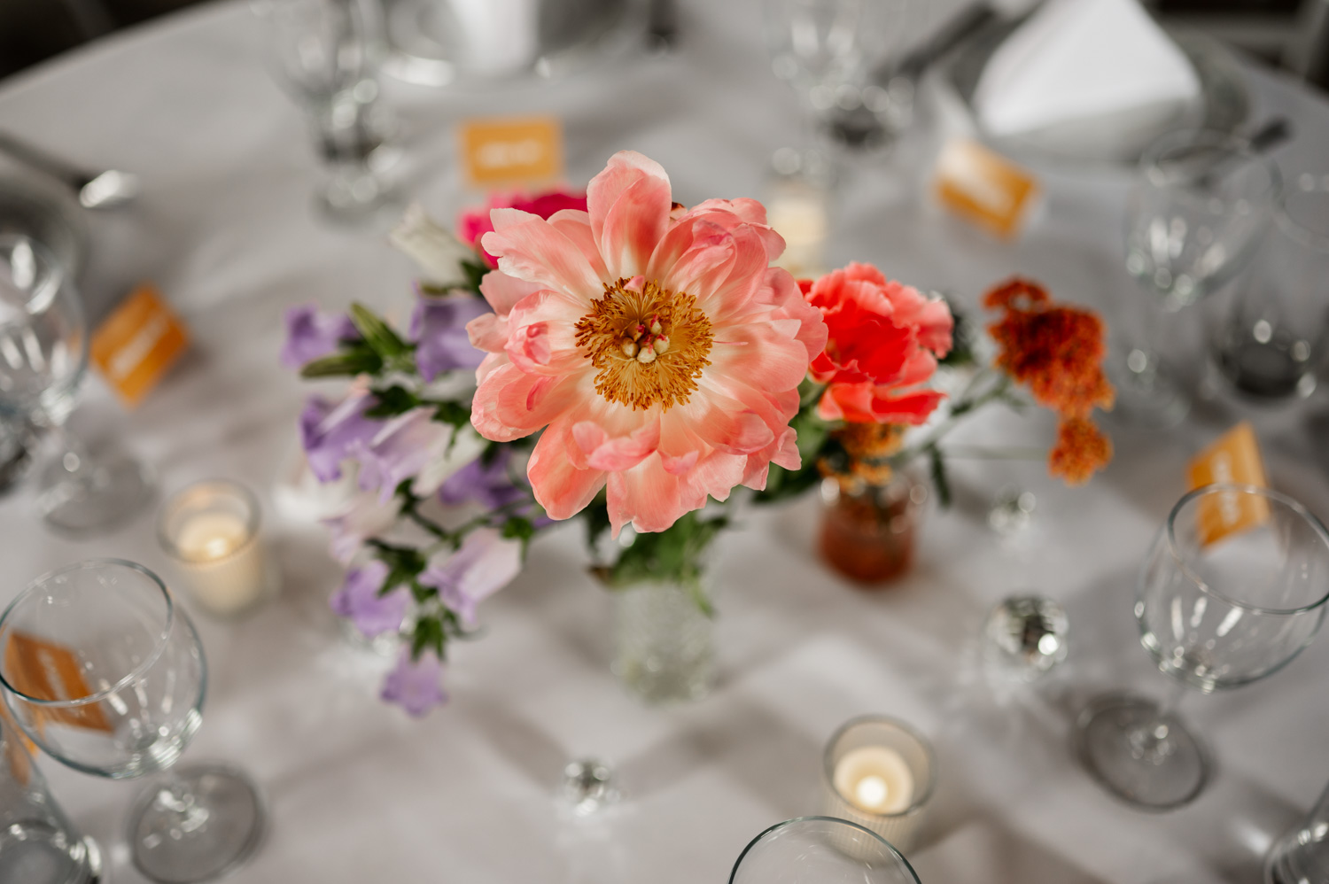 A stunning floral centerpiece with pink and purple blooms sits on the table, surrounded by empty wine glasses and lit candles, on a pristine white tablecloth.