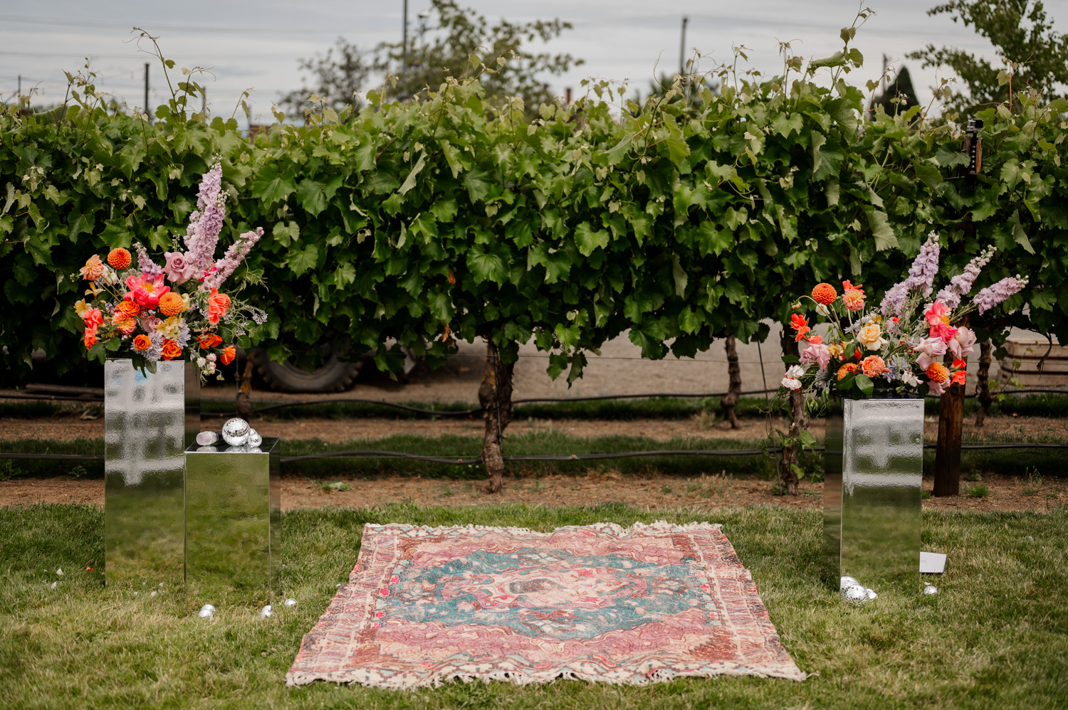 A beautifully arranged floral setup and a decorative rug are placed in front of a lush vineyard at Rossi Farms, providing a picturesque scene for wedding photos.
