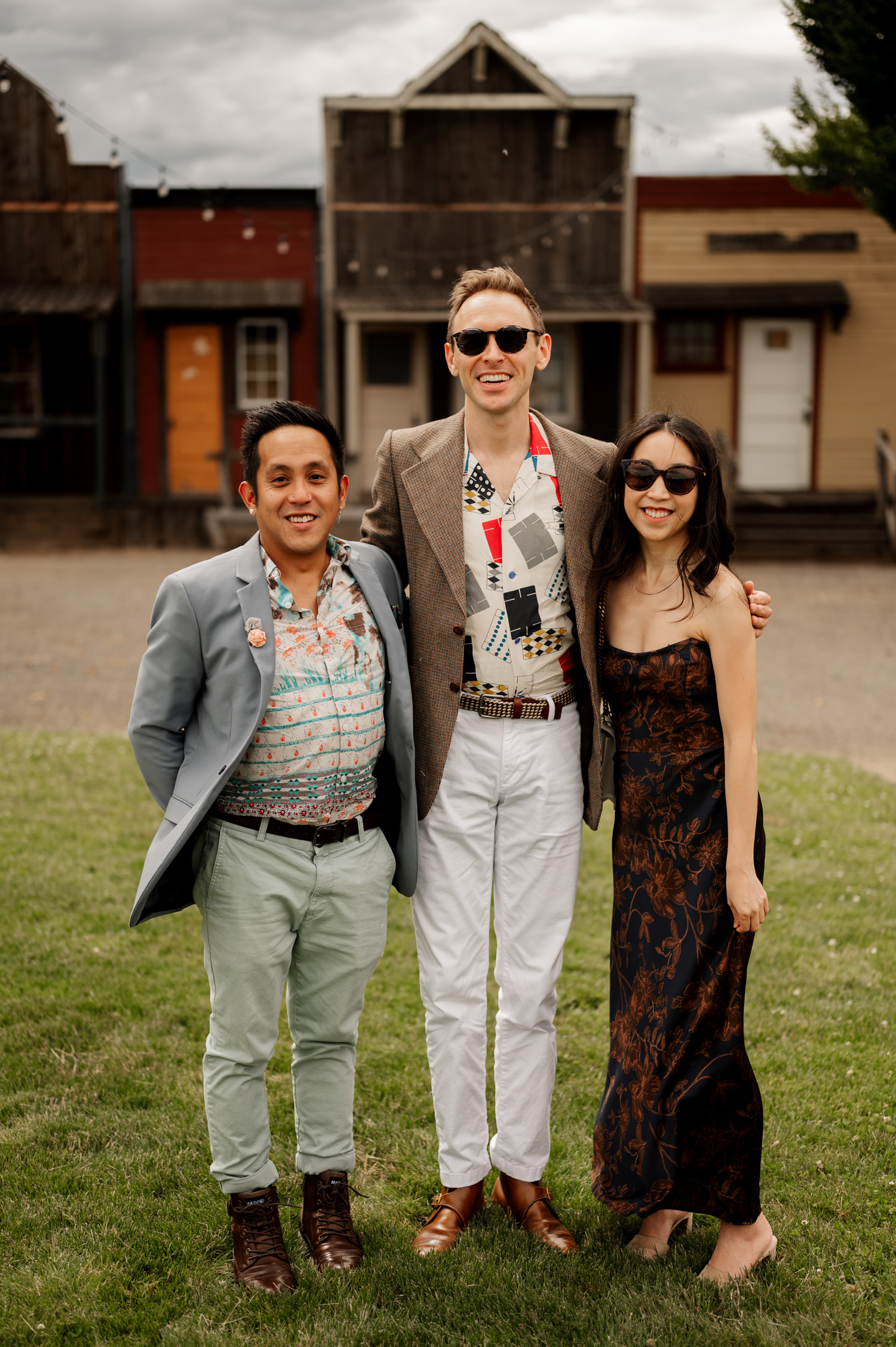 Three people stand on the grass, smiling and wearing sunglasses, with an old Western-style building in the background.