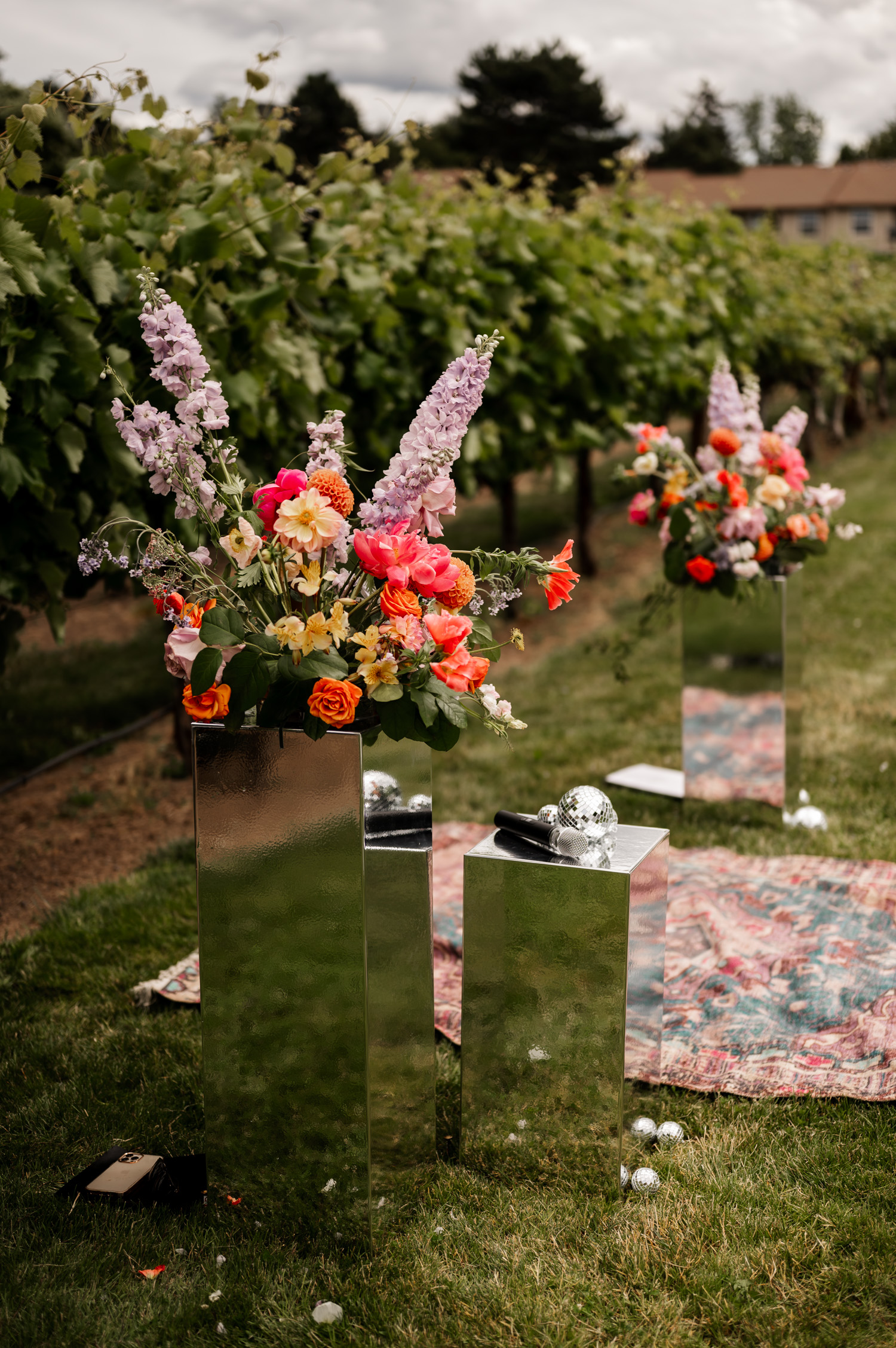Flowers in mirrored vases arranged on the grass next to a floral rug, with the vineyard of Rossi Farms visible in the background under a cloudy sky.