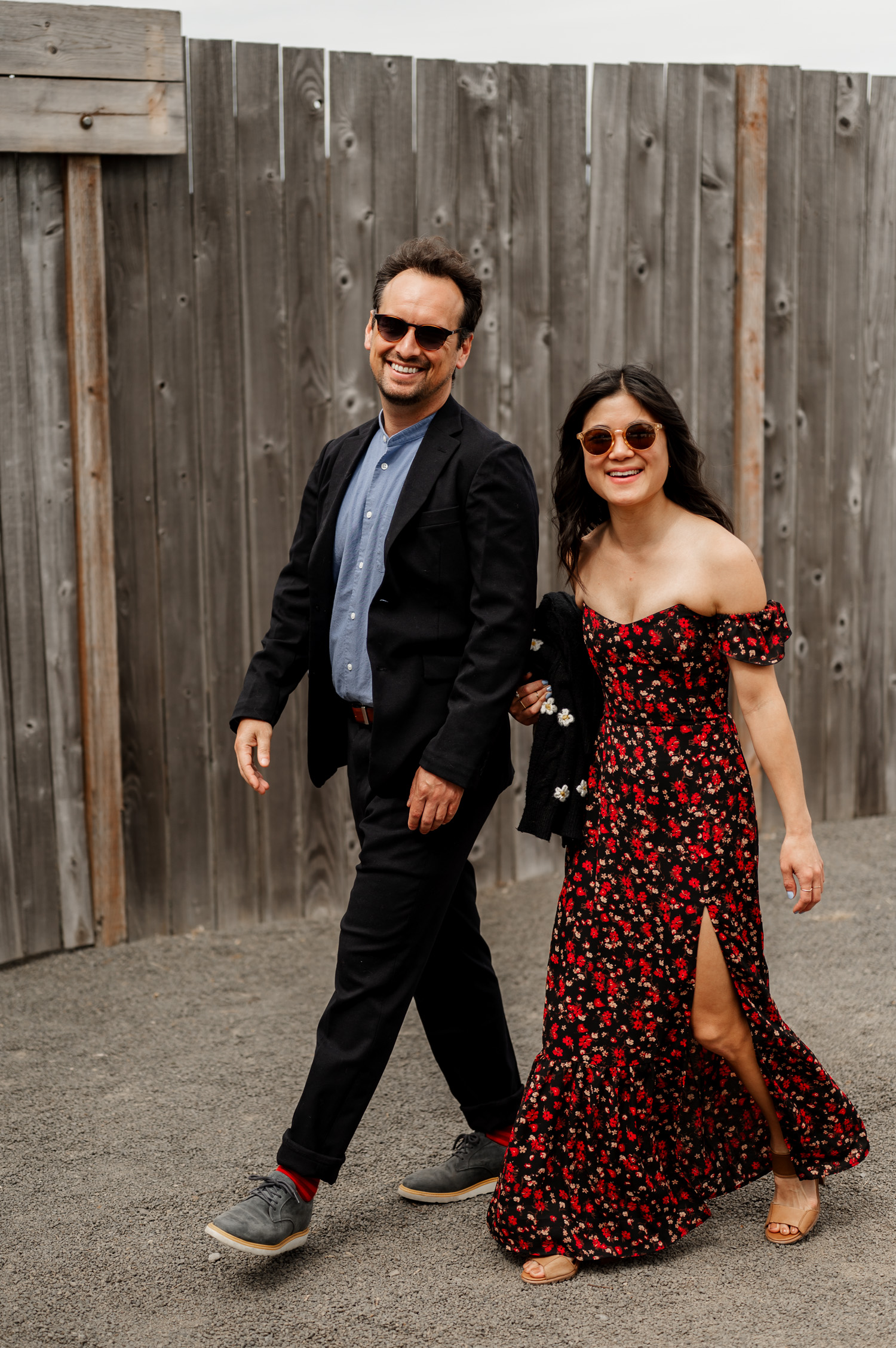 A man and woman wearing sunglasses walk joyfully in front of a wooden fence, enjoying their time at Rossi Farms during a wedding photo session.