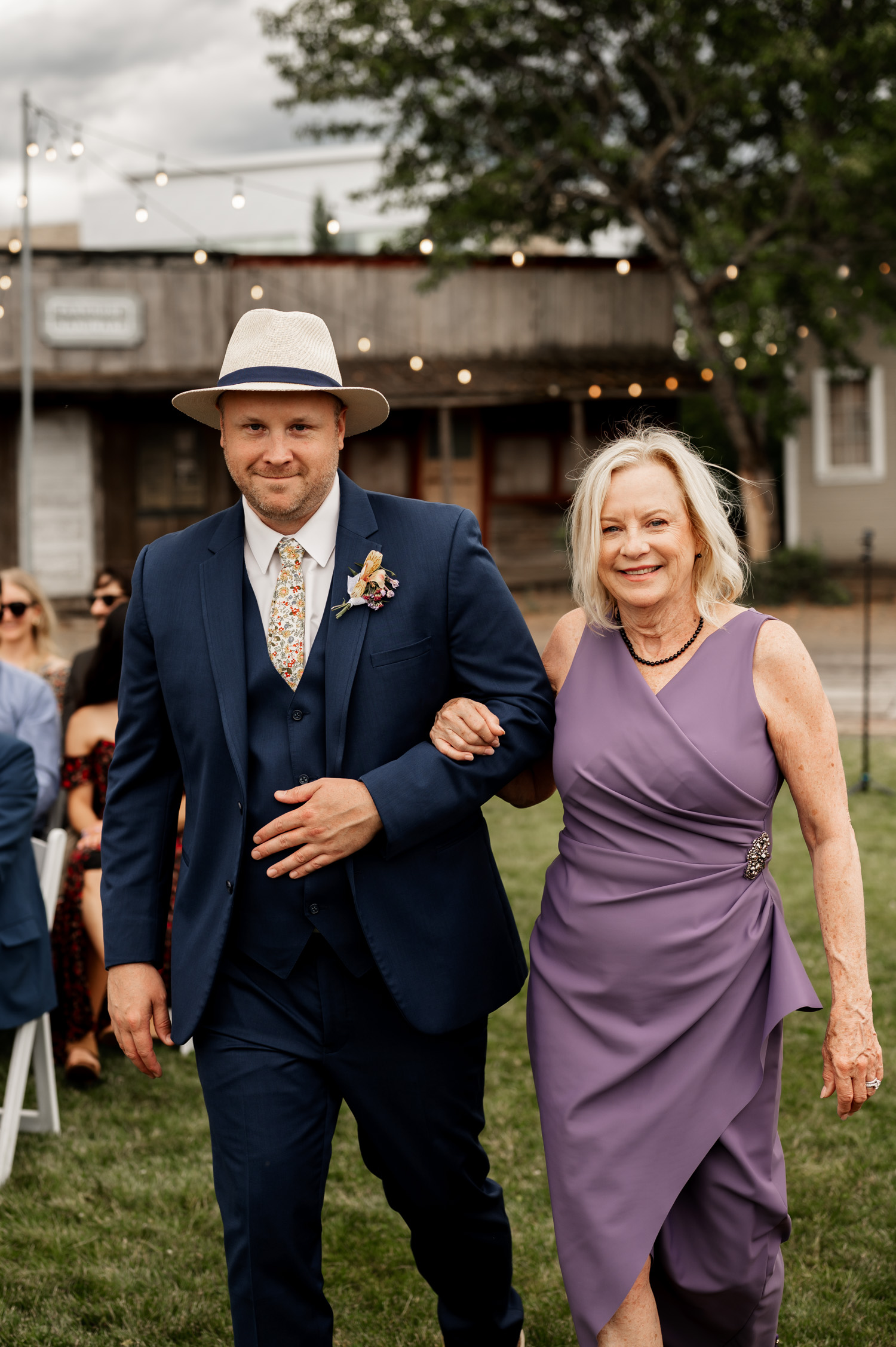 A man in a suit and hat walks arm-in-arm with a woman in a purple dress on a grassy outdoor area at Rossi Farms.