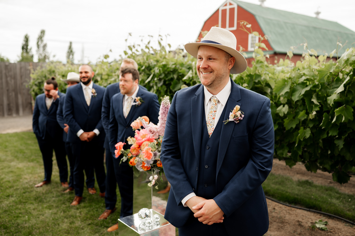 At Rossi Farms, the groom wearing a hat stands with his groomsmen dressed in blue suits outside near the vineyard, with a red barn in the background.