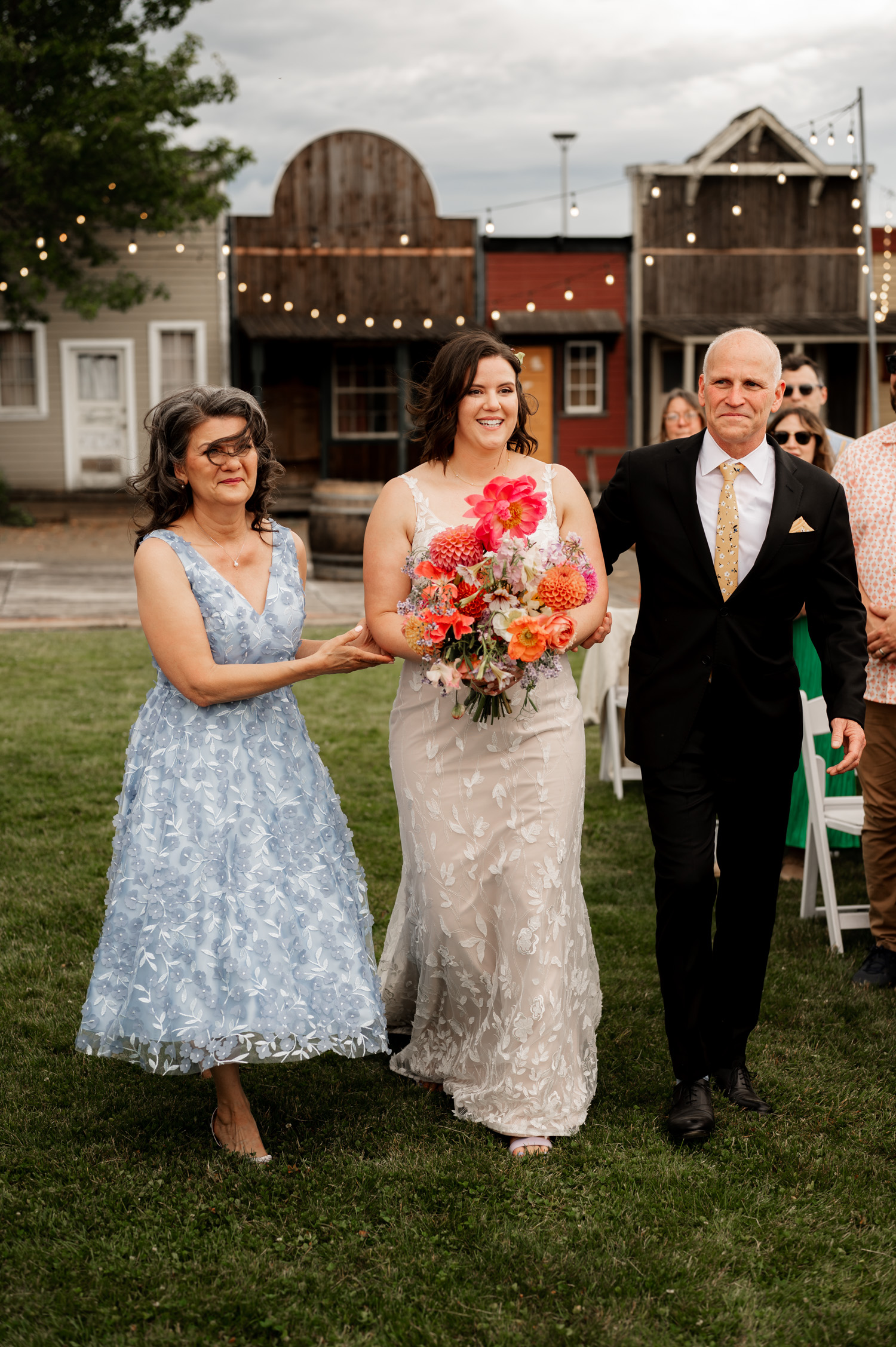 A bride in a white dress holds a bouquet while walking alongside a woman in blue and a man in a suit on the grassy path at Rossi Farms.