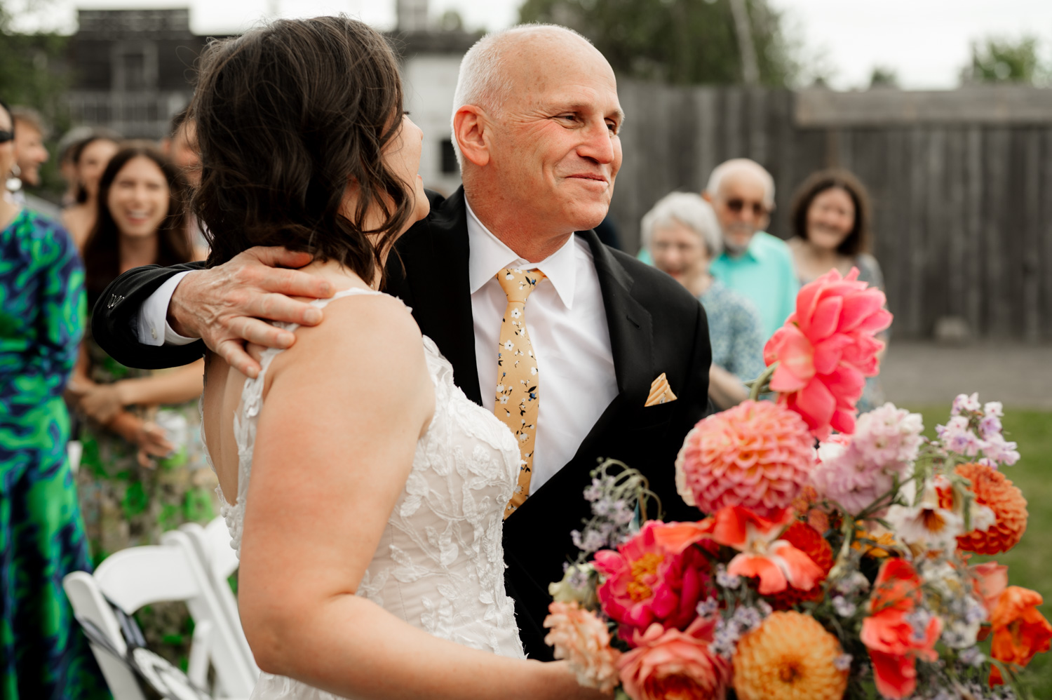 At Rossi Farms, a bride and an older man share an embrace outdoors, surrounded by smiling guests and vibrant flowers at a wedding.