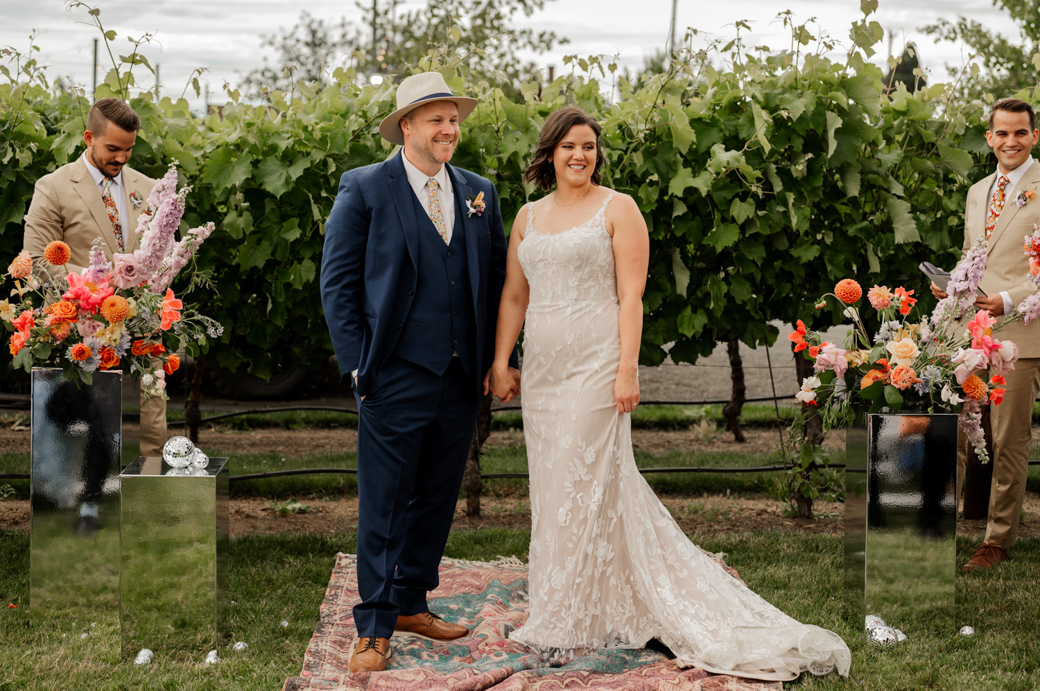 A bride and groom holding hands in a vineyard at Rossi Farms, surrounded by floral arrangements and accompanied by two groomsmen.