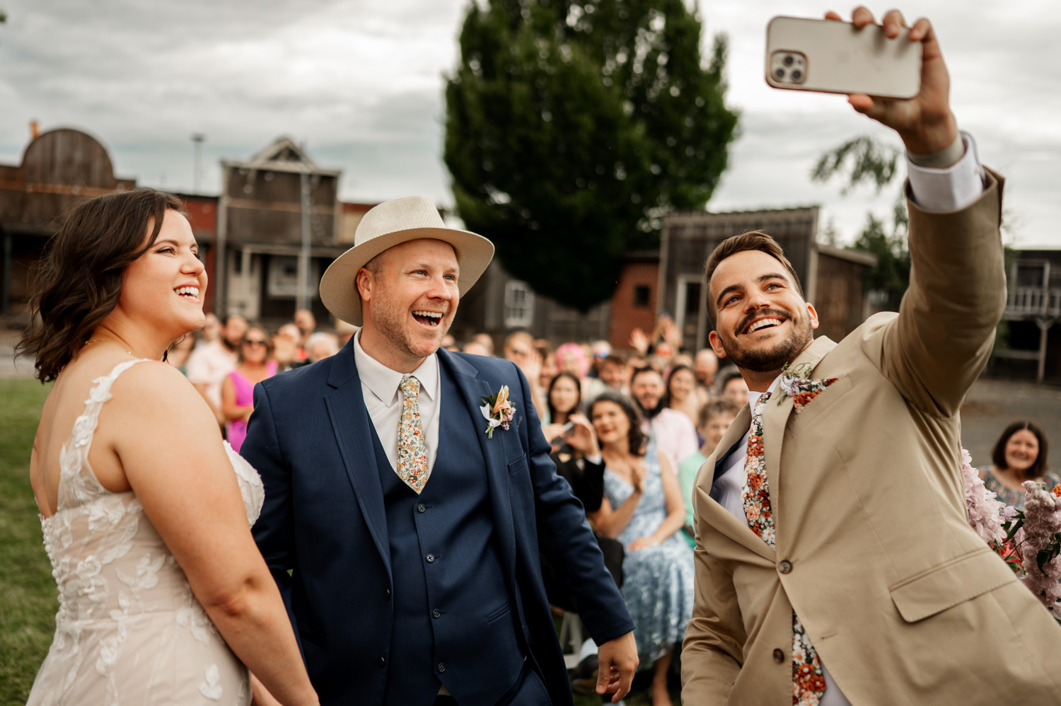 A smiling newlywed couple and a friend in suits take a selfie at Rossi Farms, with cheerful guests visible in the background.
