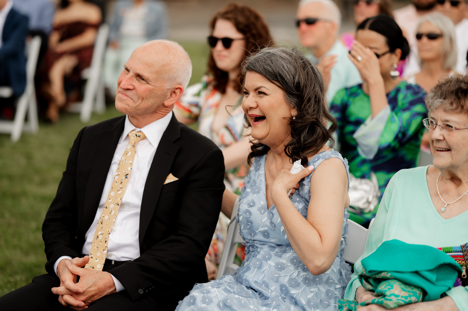 Guests seated outdoors at Rossi Farms, smiling and chatting during a wedding celebration. A woman in a blue dress stands out with her joyful expression, contributing to the lively atmosphere captured in the photos.