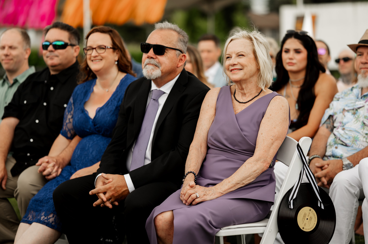 People sitting outdoors, smiling and dressed up at an event at Rossi Farms. A woman in a purple outfit and a man in a suit are seated in the front row, enjoying the joyful atmosphere of wedding photos.