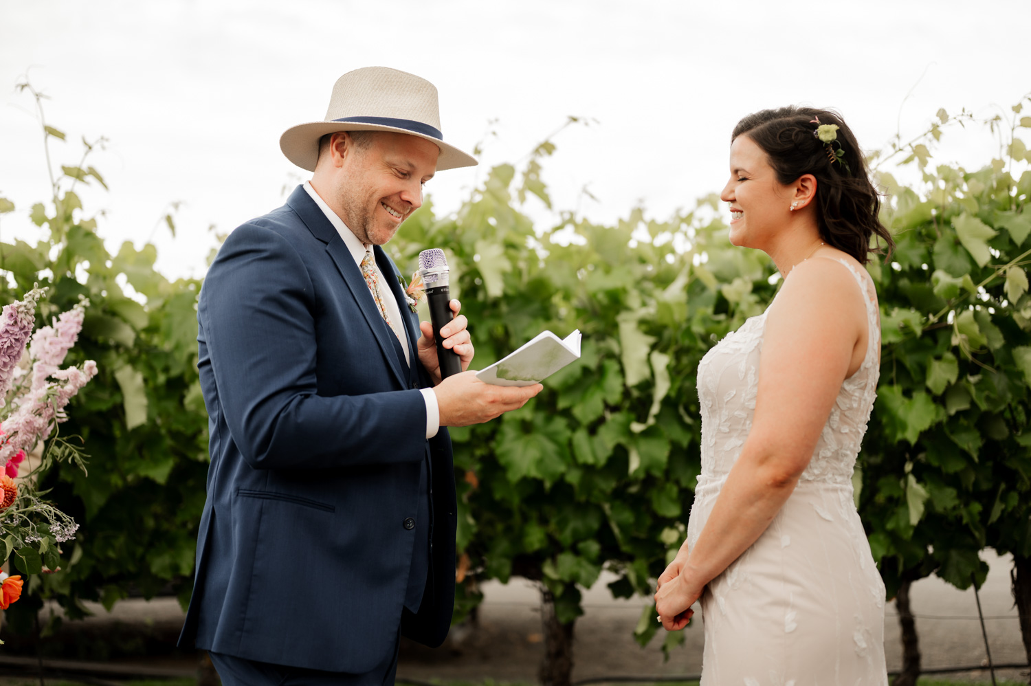 A man in a hat reads from a notebook and smiles at a woman in a white dress. They are standing against the backdrop of picturesque vineyards at Rossi Farms.