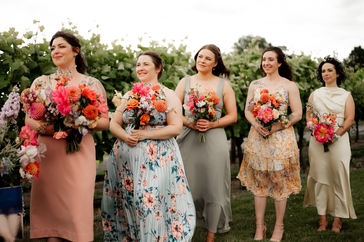 Five women in colorful dresses hold vibrant flower bouquets, standing together outdoors with greenery in the background.