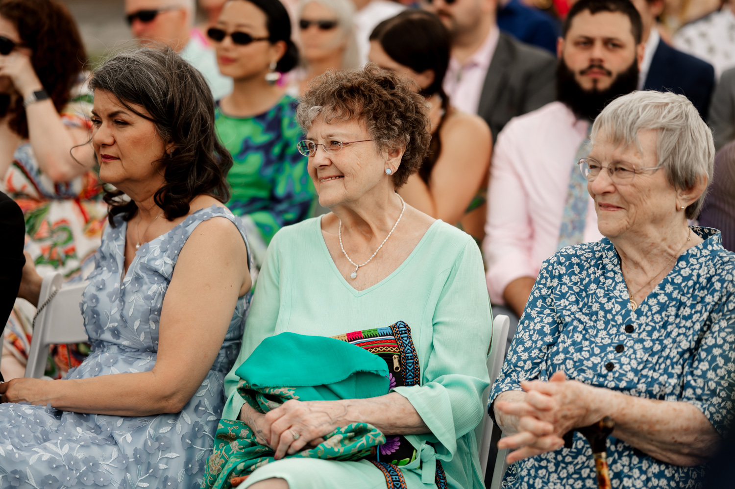 Three women sitting outdoors at an event, surrounded by other attendees, smiling and enjoying the lively atmosphere at Rossi Farms.