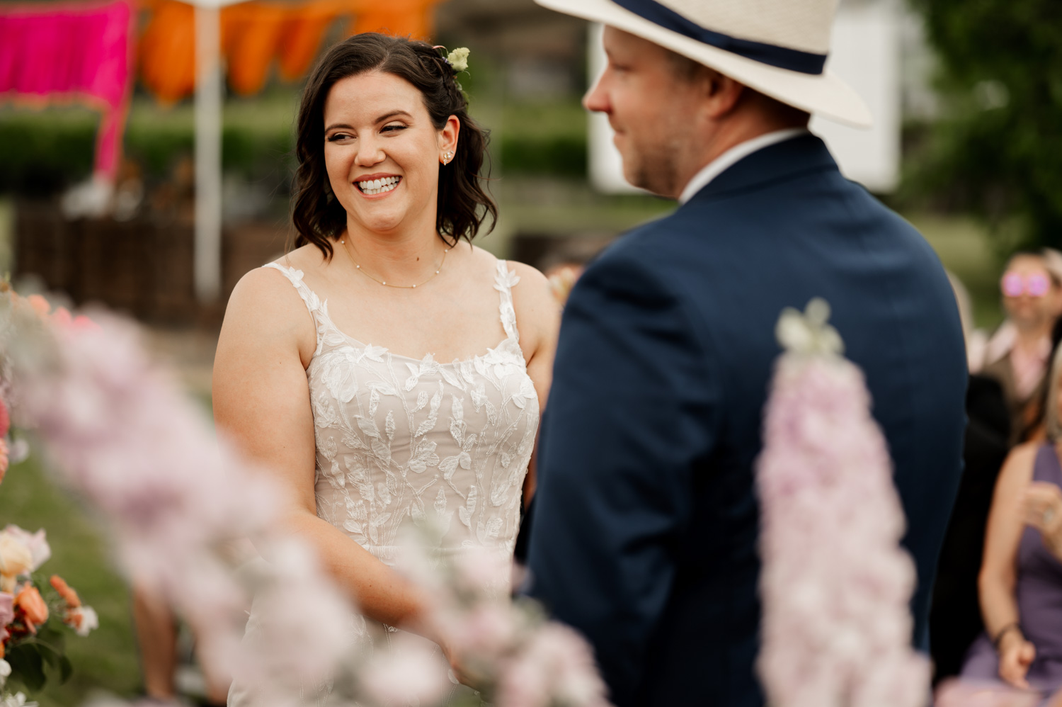 A bride smiling at her groom during an outdoor ceremony at Rossi Farms, with colorful decorations in the background.