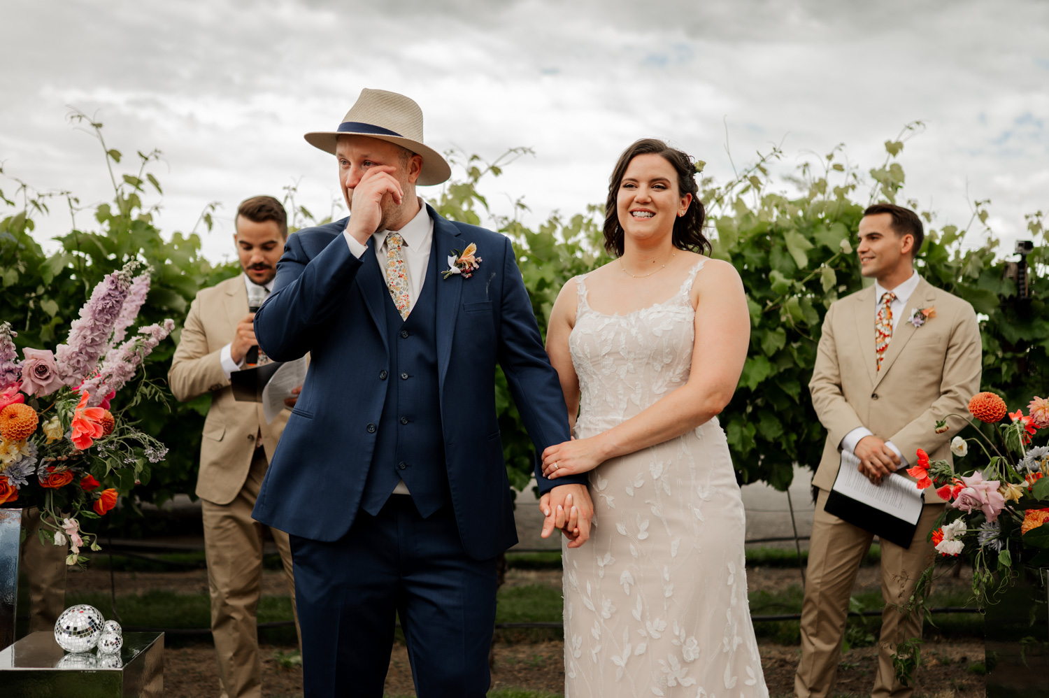 At a Rossi Farms wedding, the bride and groom stand outdoors with joyful expressions, surrounded by two groomsmen and vibrant floral arrangements.