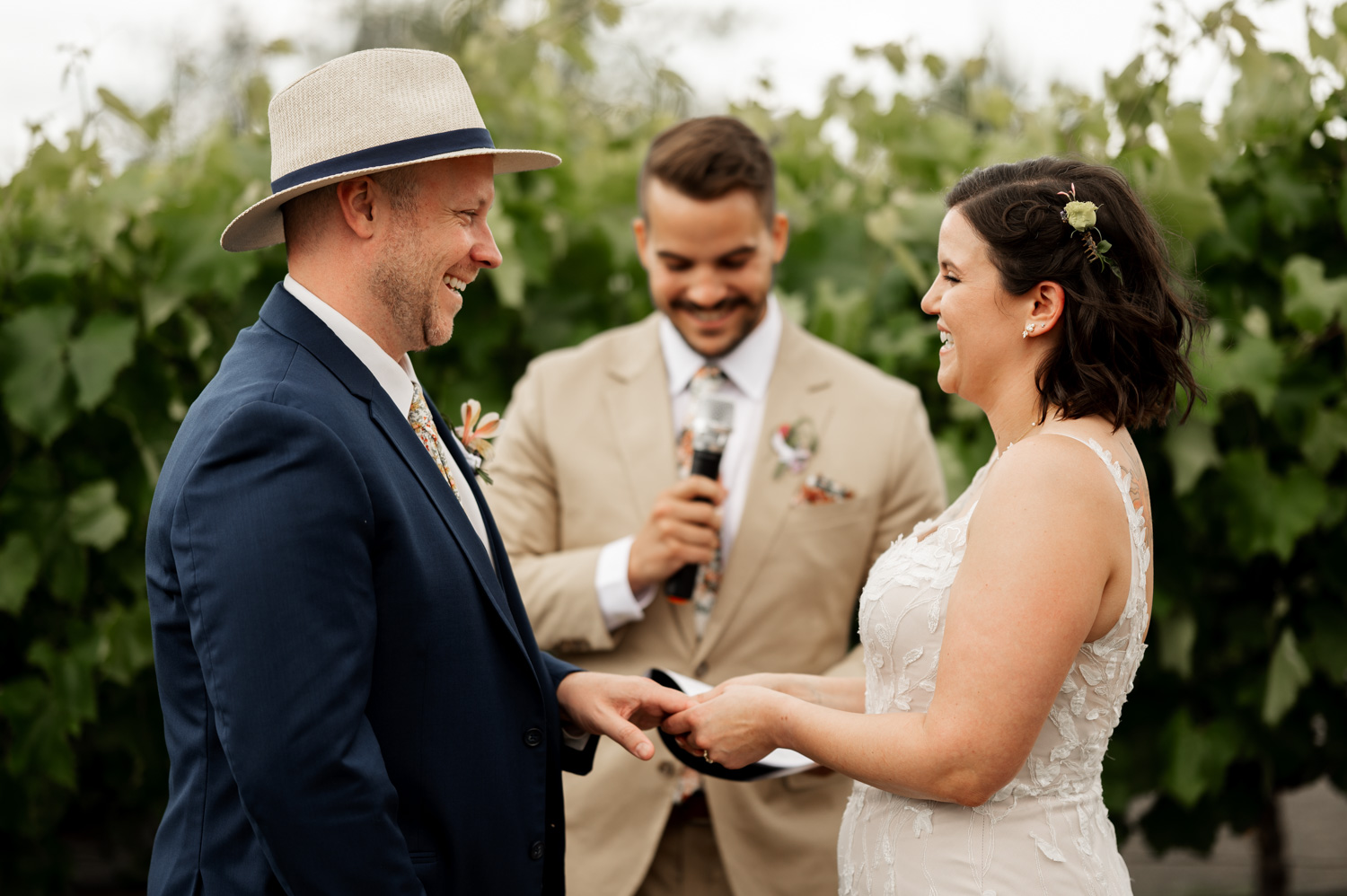 A couple holds hands and exchanges vows outdoors at Rossi Farms, with a smiling officiant standing in front of a lush green backdrop.