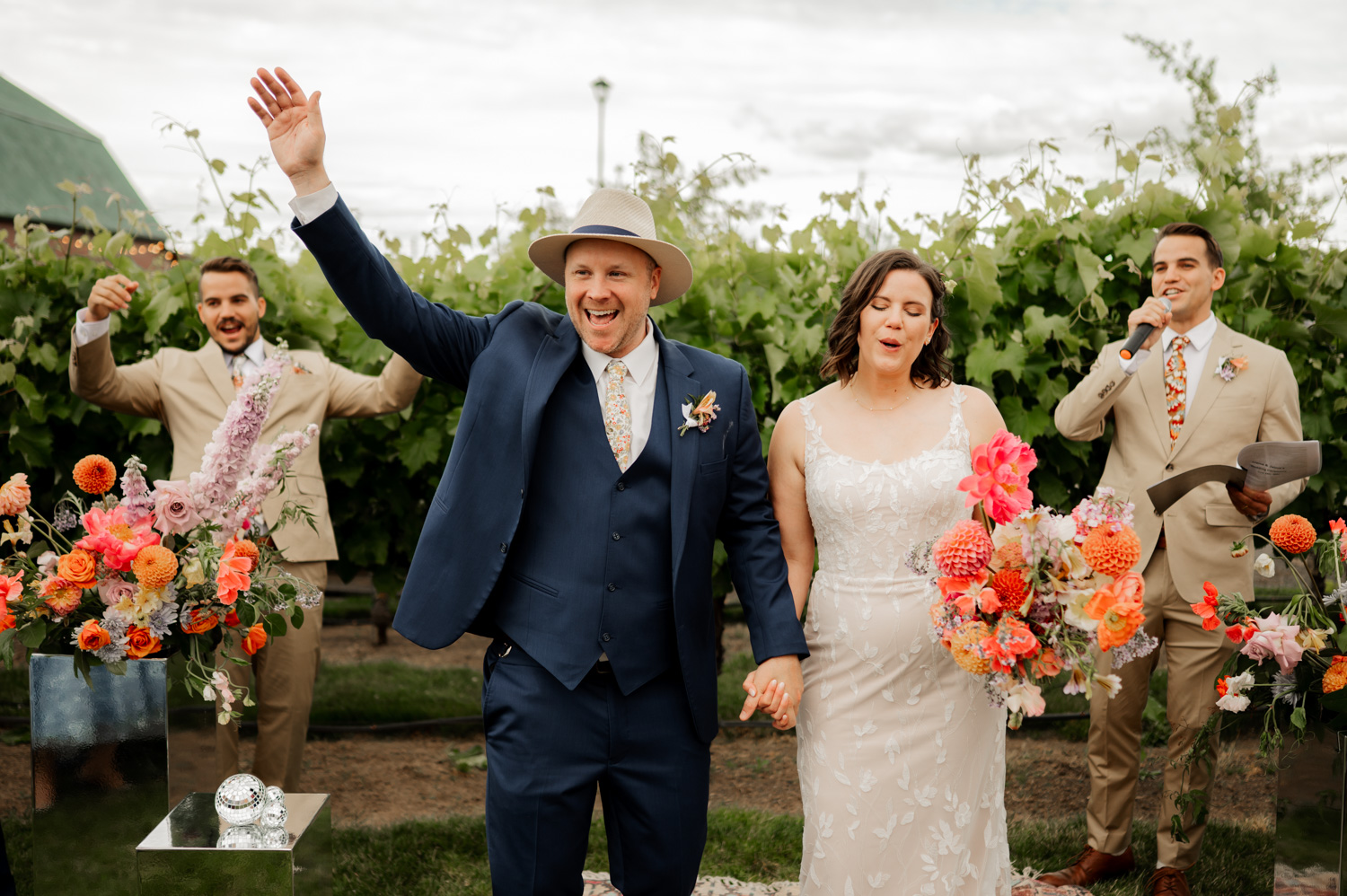 Bride and groom celebrating joyfully outside at Rossi Farms, surrounded by lush greenery and vibrant flowers, with two groomsmen cheering.
