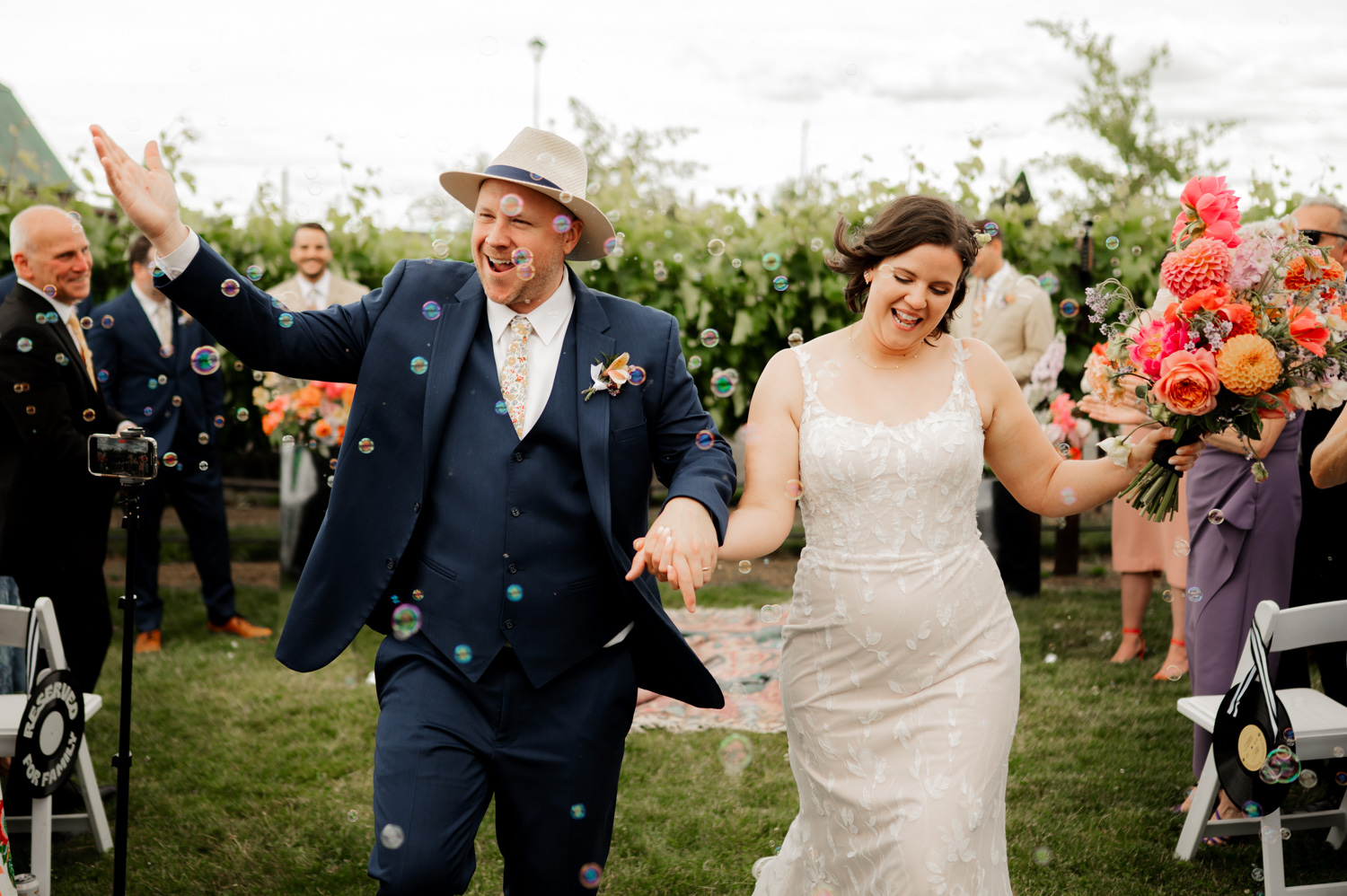 Newlyweds walking hand in hand through floating bubbles at Rossi Farms, with the bride holding colorful flowers and happy guests looking on.
