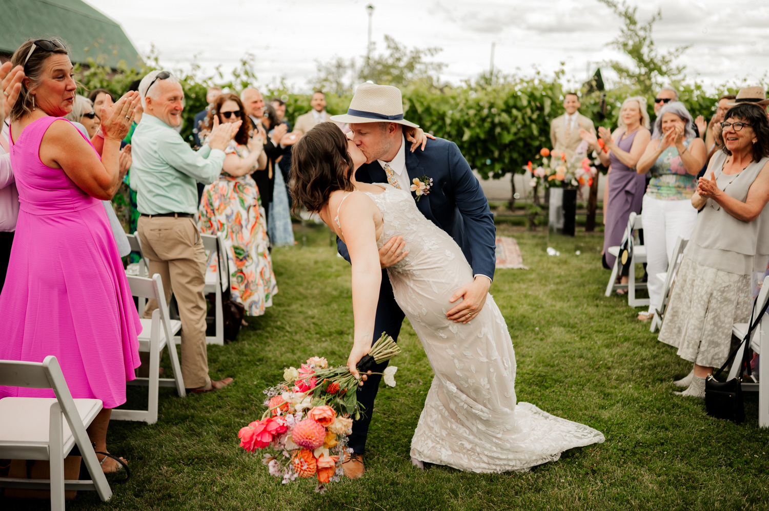 Bride and groom share a kiss at their Rossi Farms wedding as guests applaud, with lush greenery in the background.