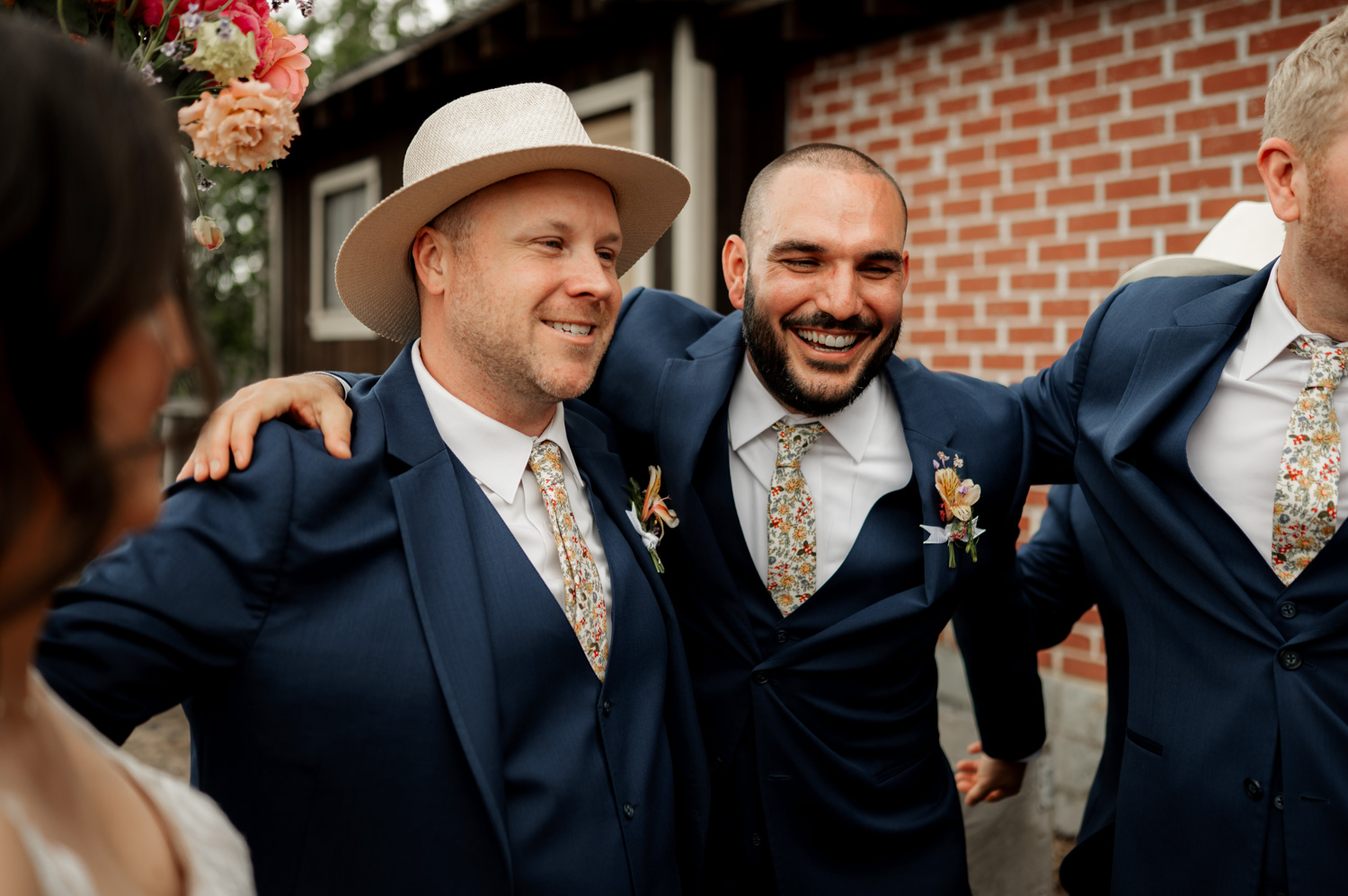 Two smiling groomsmen in blue suits, one wearing a hat, stand arm-in-arm outside a brick building at Rossi Farms wedding.