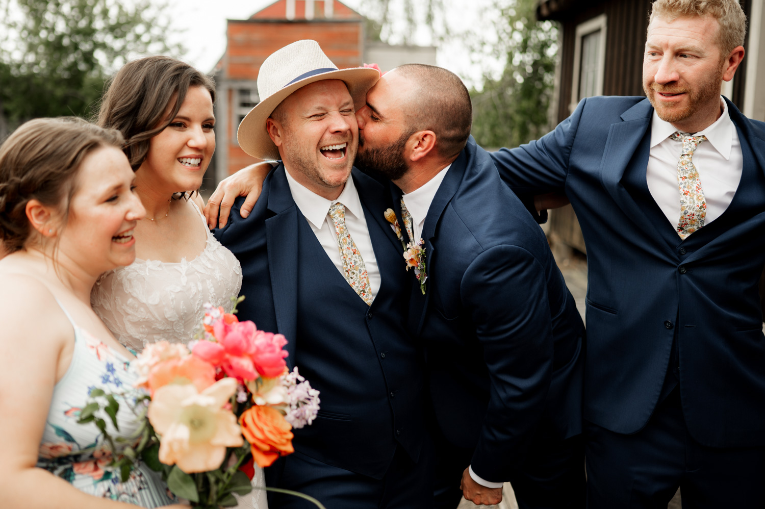 Five people in formal attire smile at a Rossi Farms wedding. A man kisses another mans cheek, and a woman holds a colorful bouquet.