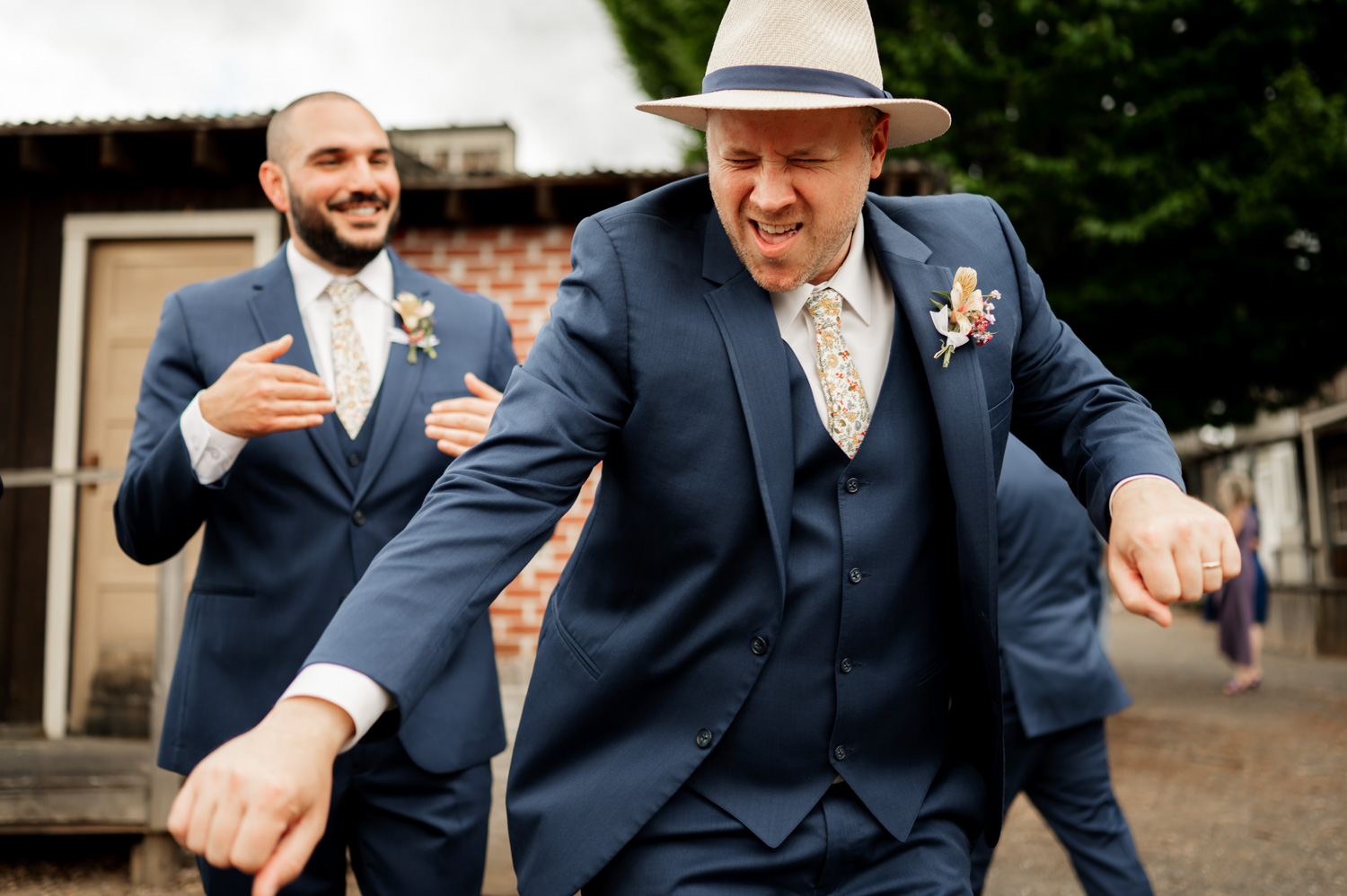 Two men in blue suits dance joyfully outdoors at Rossi Farms; one wears a hat and boutonniere, while the other claps and smiles radiantly, capturing the delight of wedding photos.