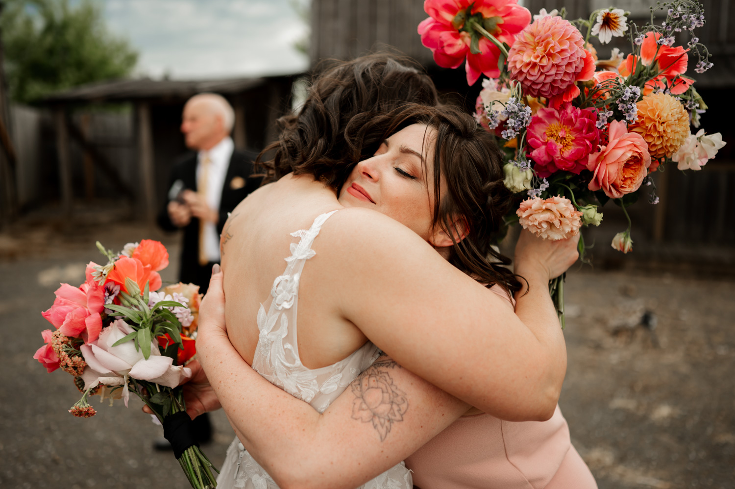 Two women embrace, each holding vibrant bouquets, with a man in a suit standing gracefully in the background at the Rossi Farms wedding venue.