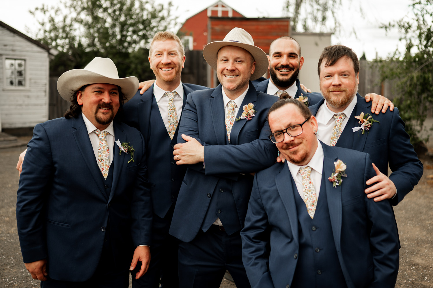 Six men in blue suits and patterned ties, smiling as they pose outdoors at Rossi Farms; two are wearing stylish hats, capturing a joyful moment.