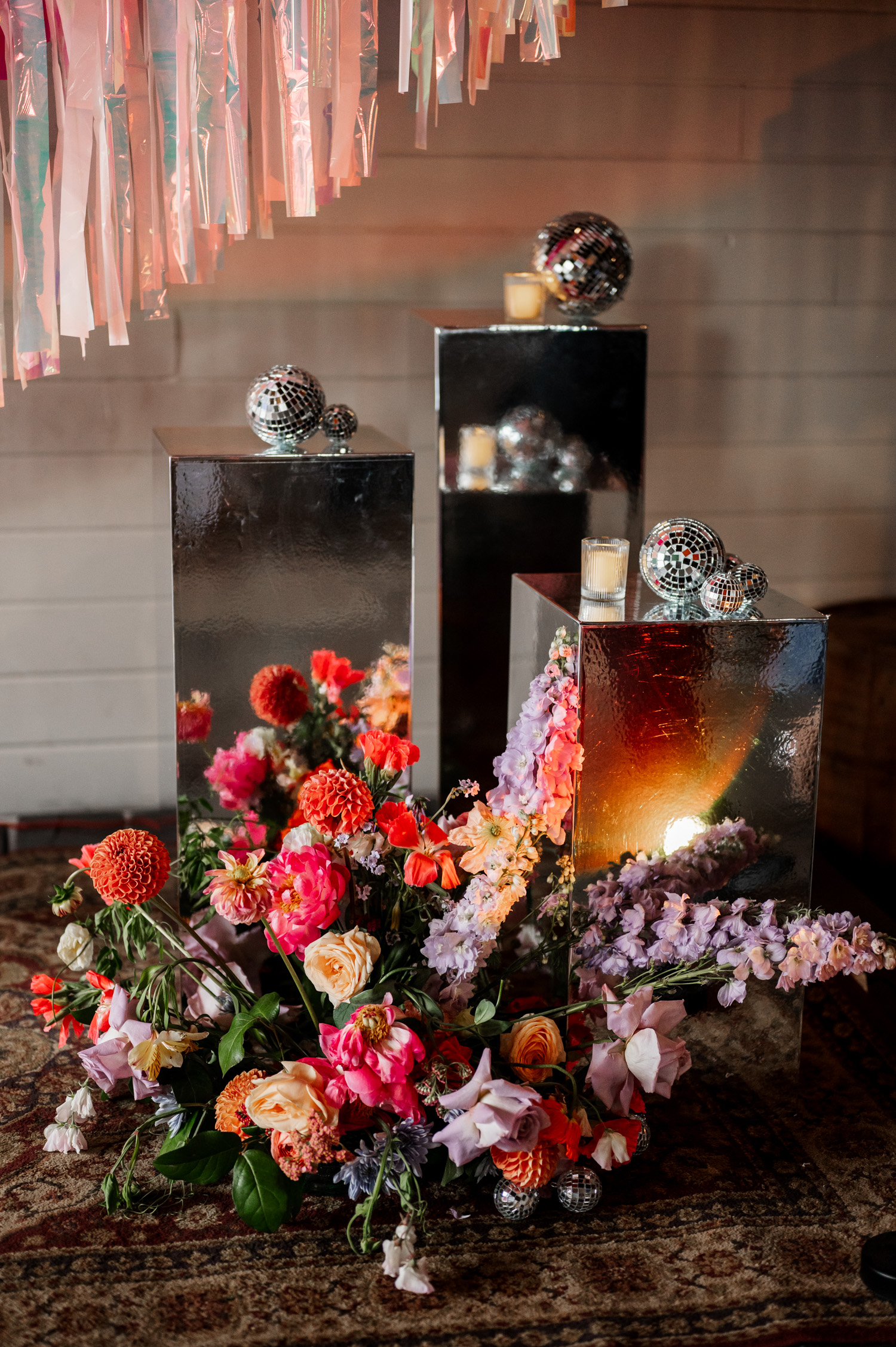Floral arrangement featuring vibrant blooms alongside mirrored pedestals, disco balls, and candles at a Rossi Farms wedding. The scene is set under hanging ribbons.